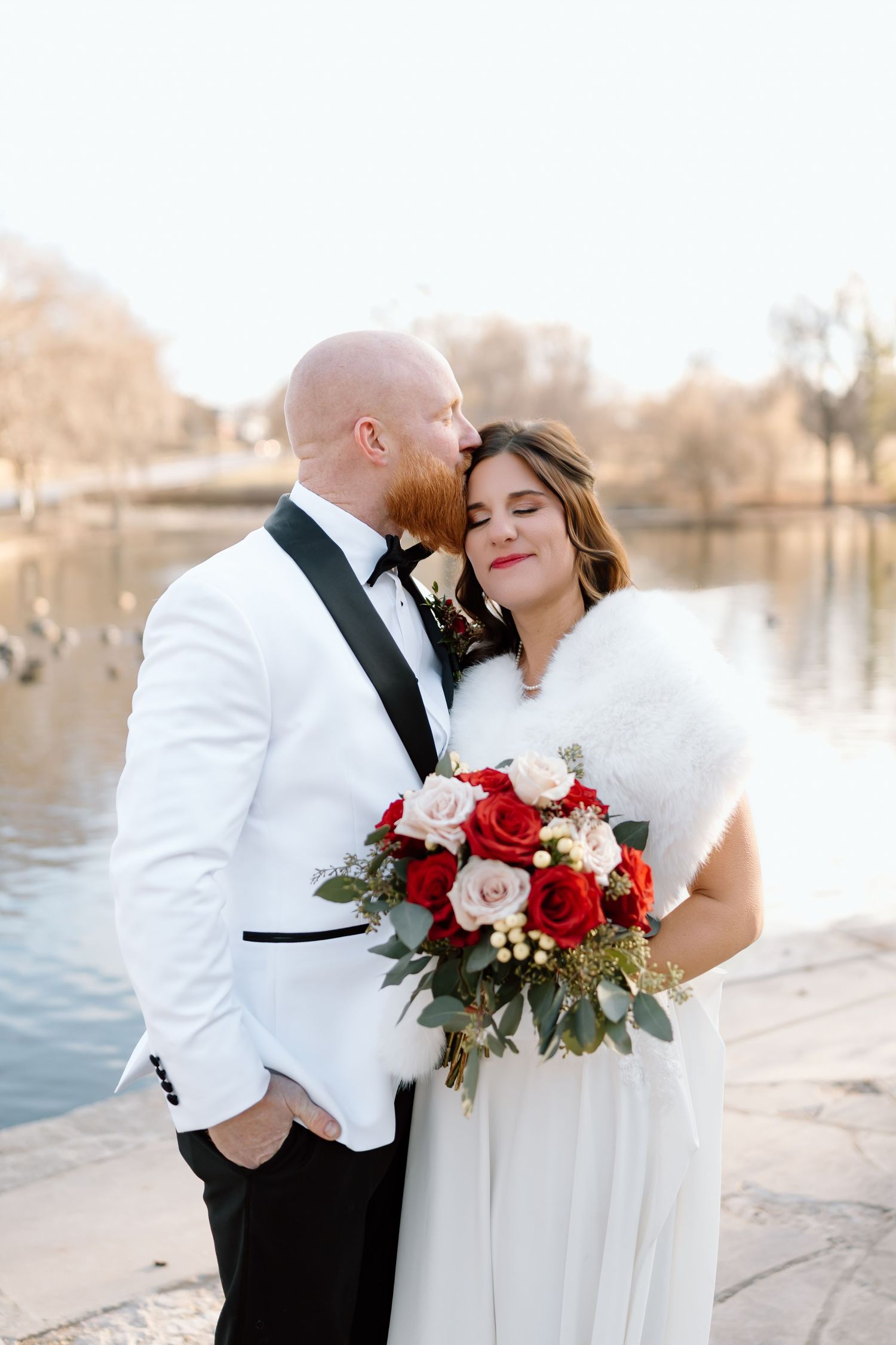 Romantic winter wedding couple embraces by a lake with a red and white rose bouquet at sunset.
