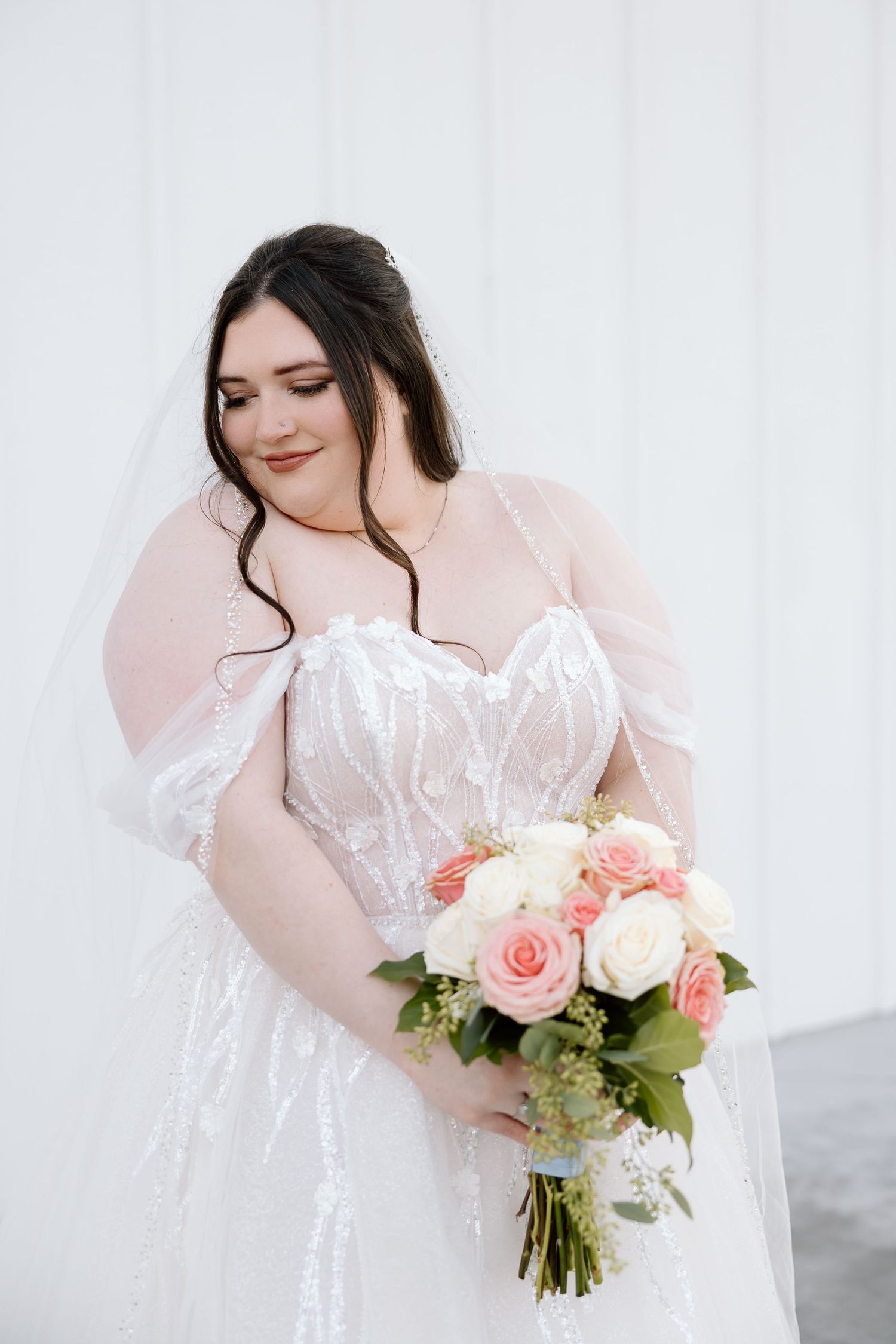 Bride in lace and tulle gown holding pink and white rose bouquet against white backdrop.