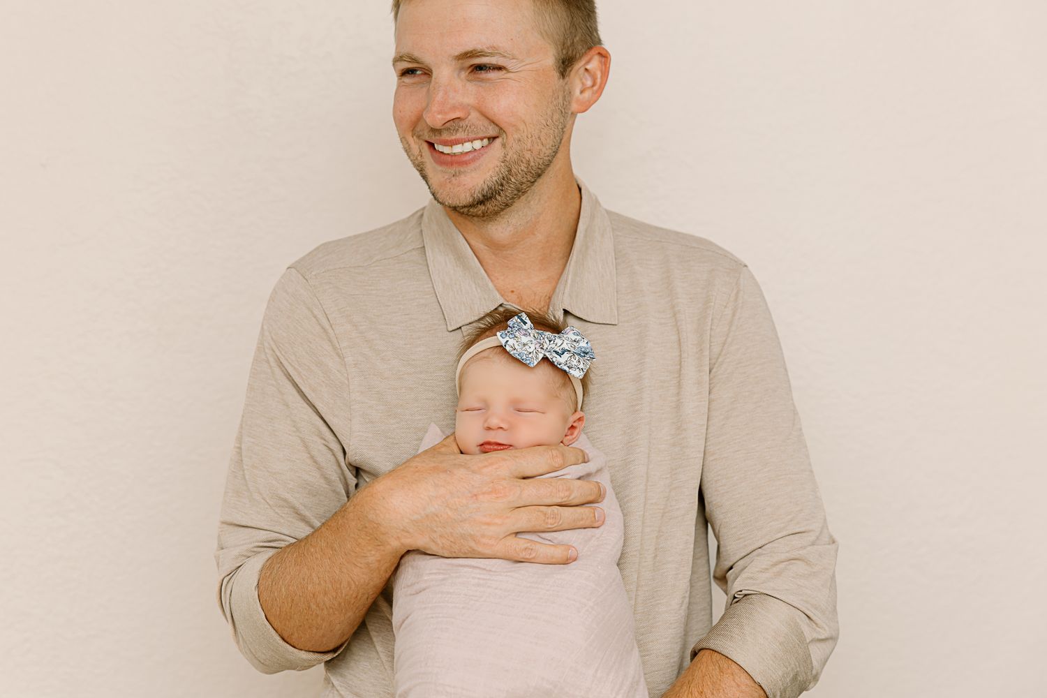 Adult holding sleepy newborn wearing sparkly bow against neutral backdrop.