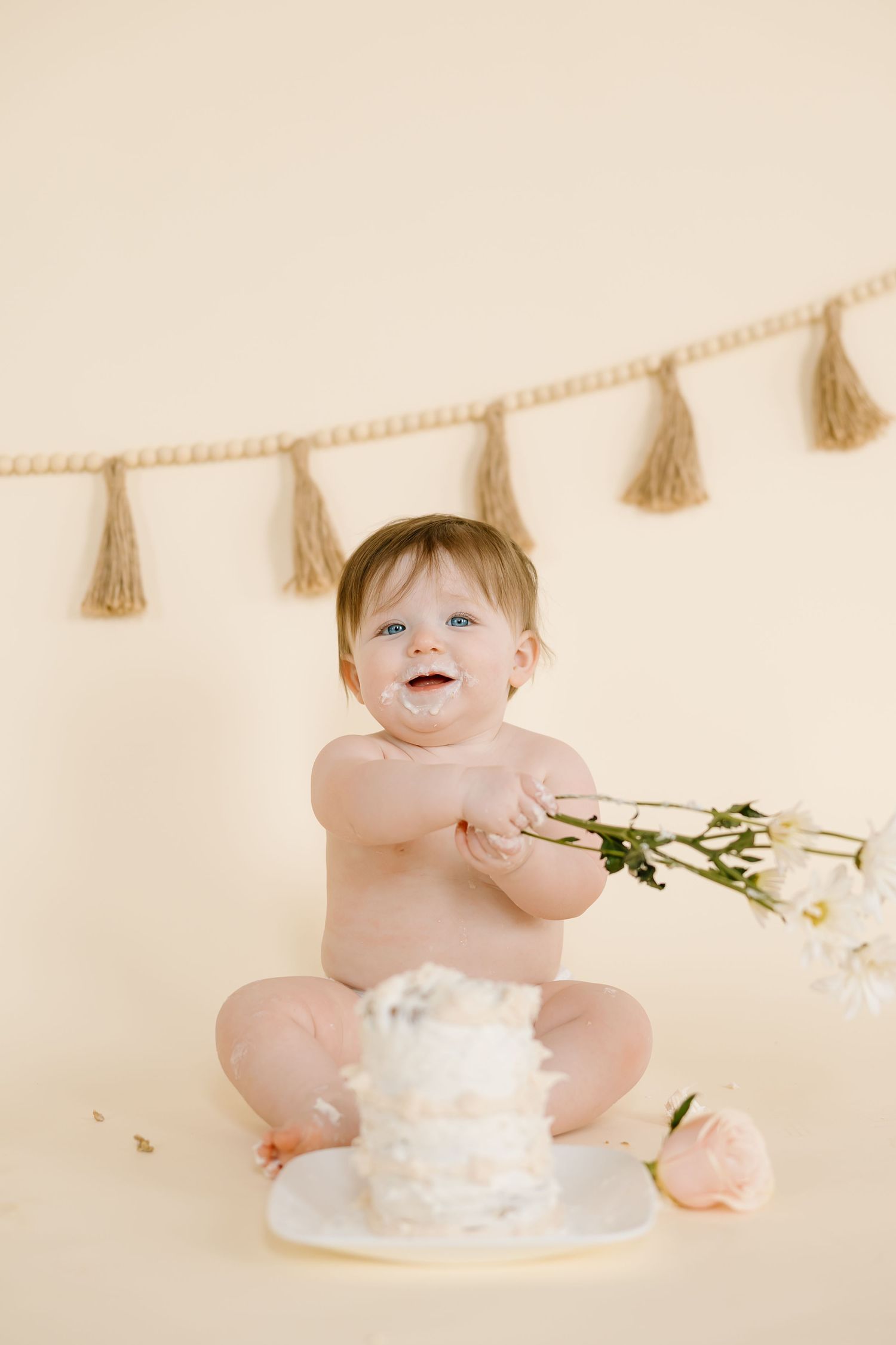 Happy baby holding flowers near white cake against beige wall with tassel garland.