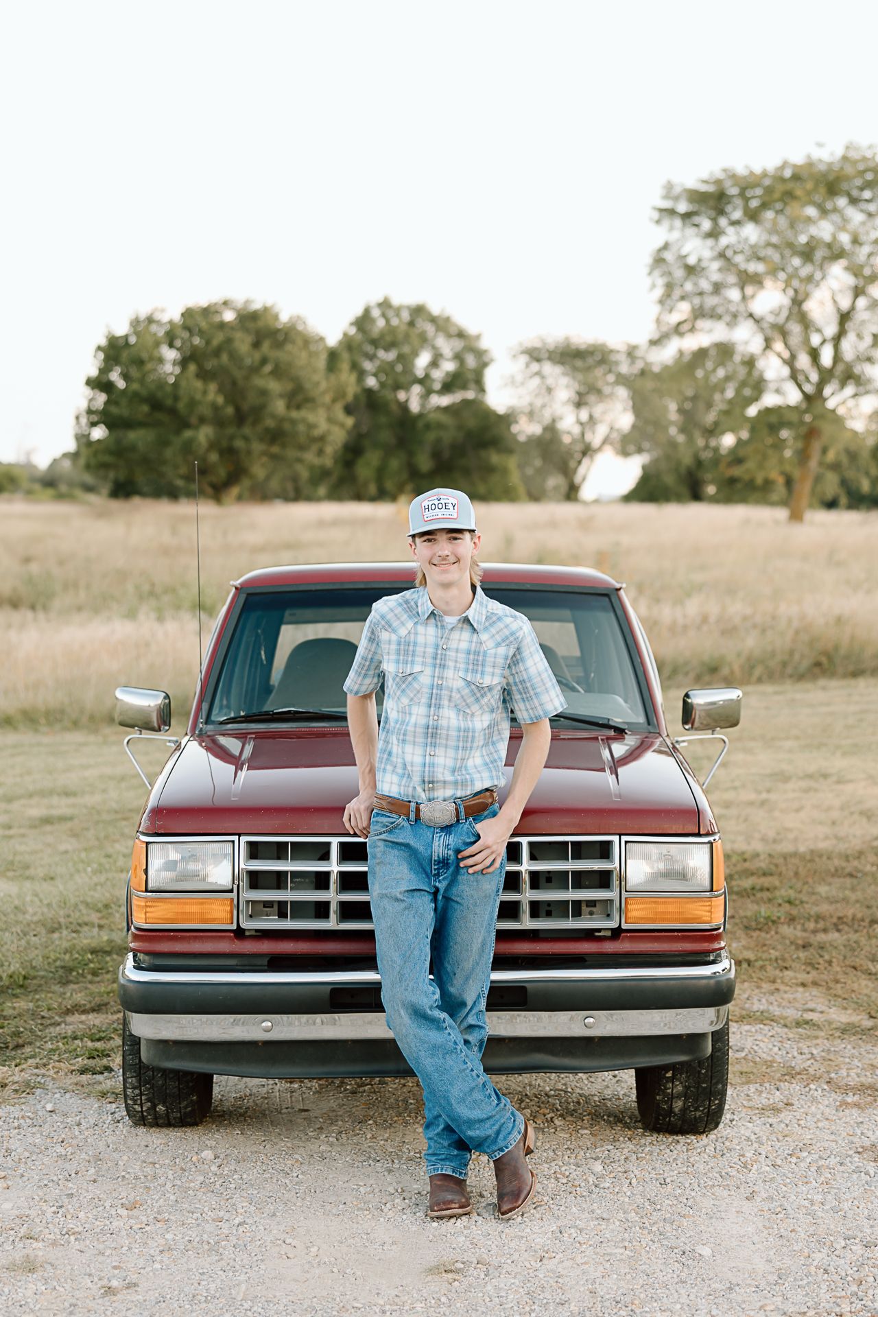 Someone in western wear leaning against the front of a vintage Ford pickup truck in a grassy field.