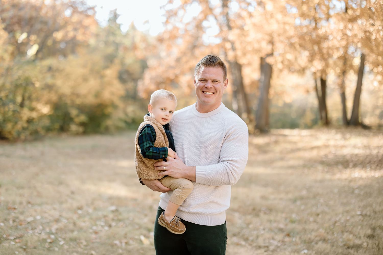 Joyful parental moment captured outdoors with fall trees and soft lighting in the background.