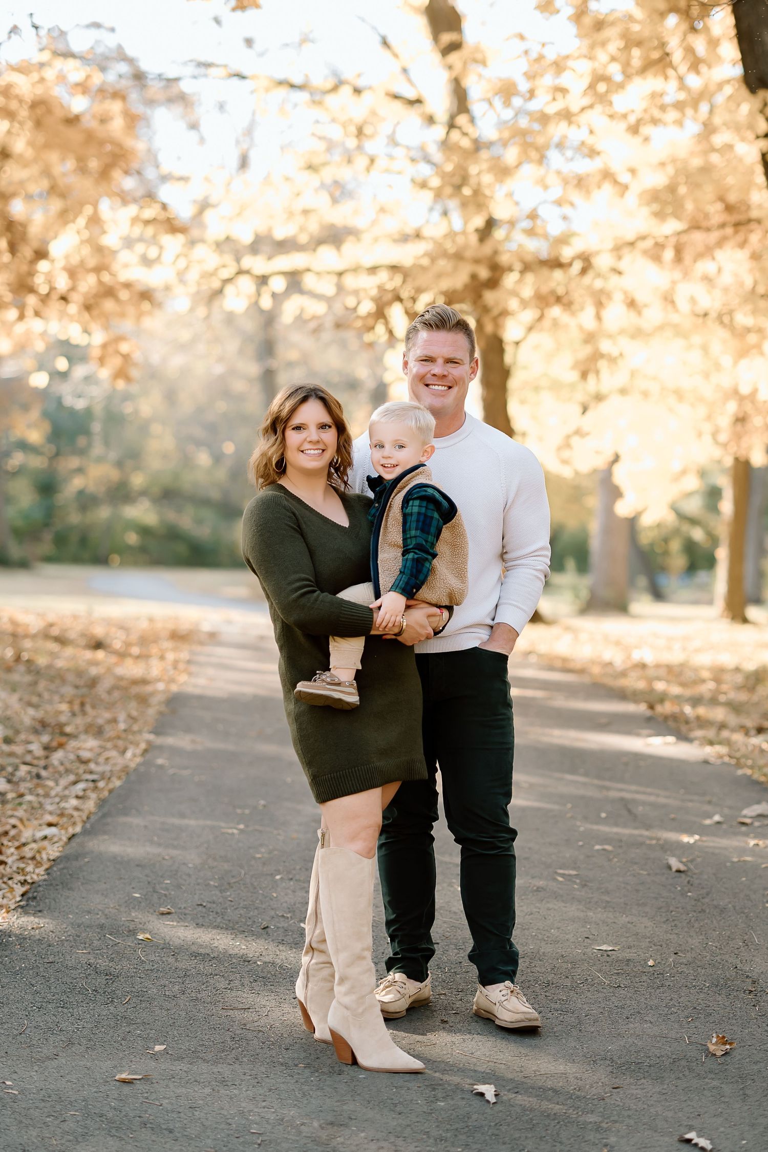A couple and child pose together on a park path surrounded by golden autumn trees.
