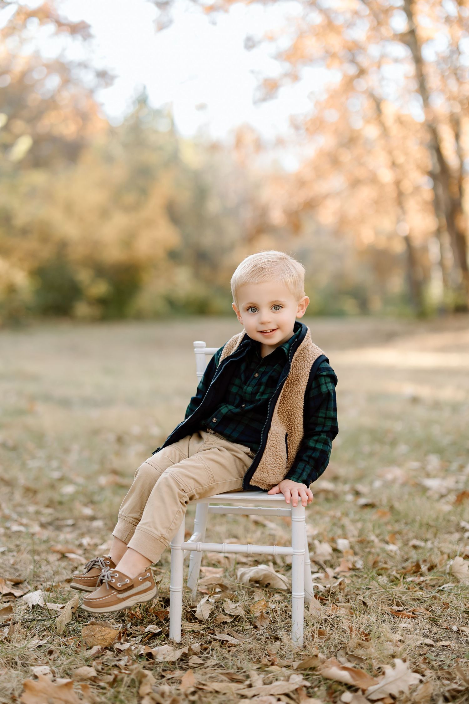 Child wearing plaid shirt and vest sits on white chair surrounded by fall foliage.