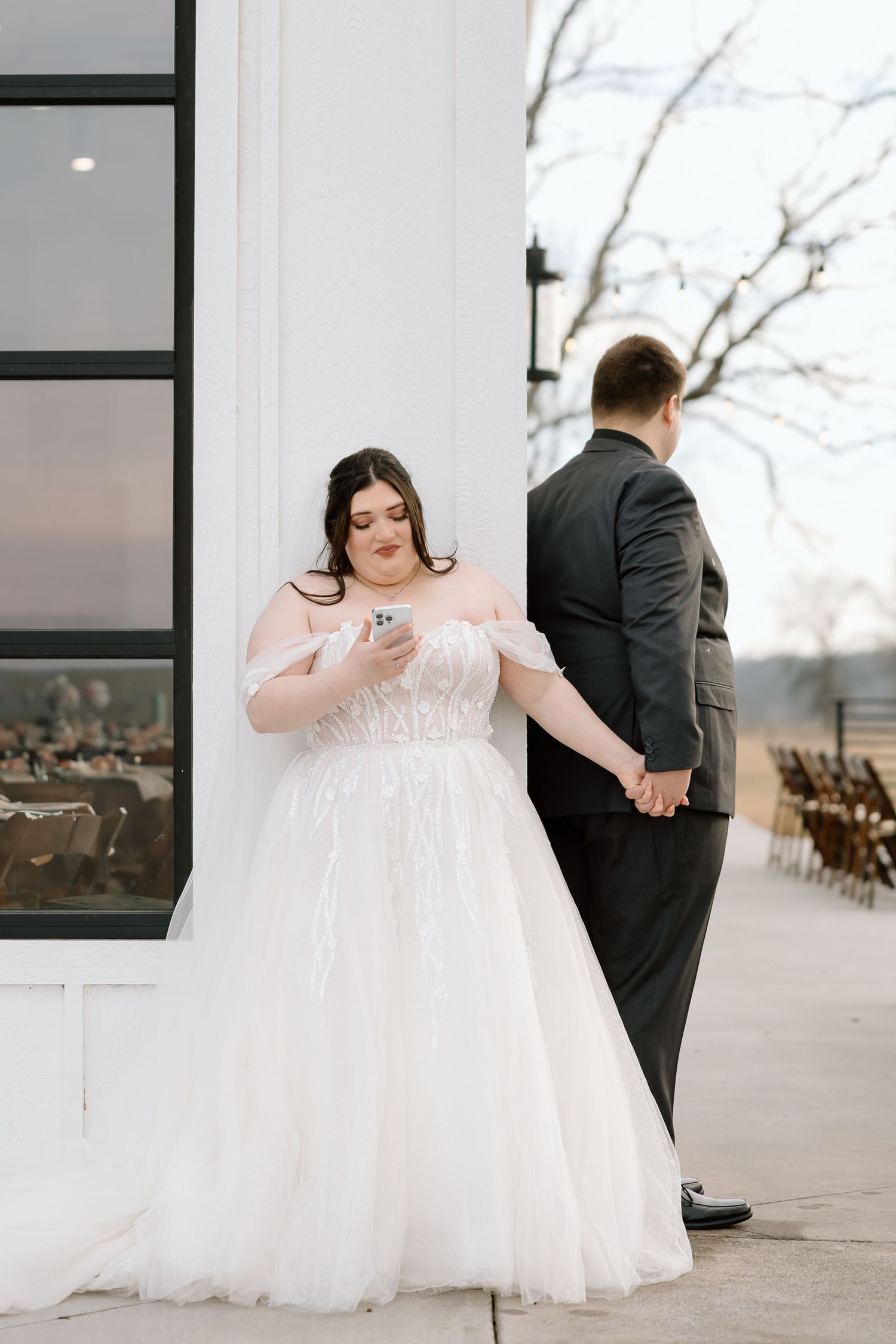 Bride in flowing white gown and groom in dark suit share a first look moment on a patio with rustic chairs in background.