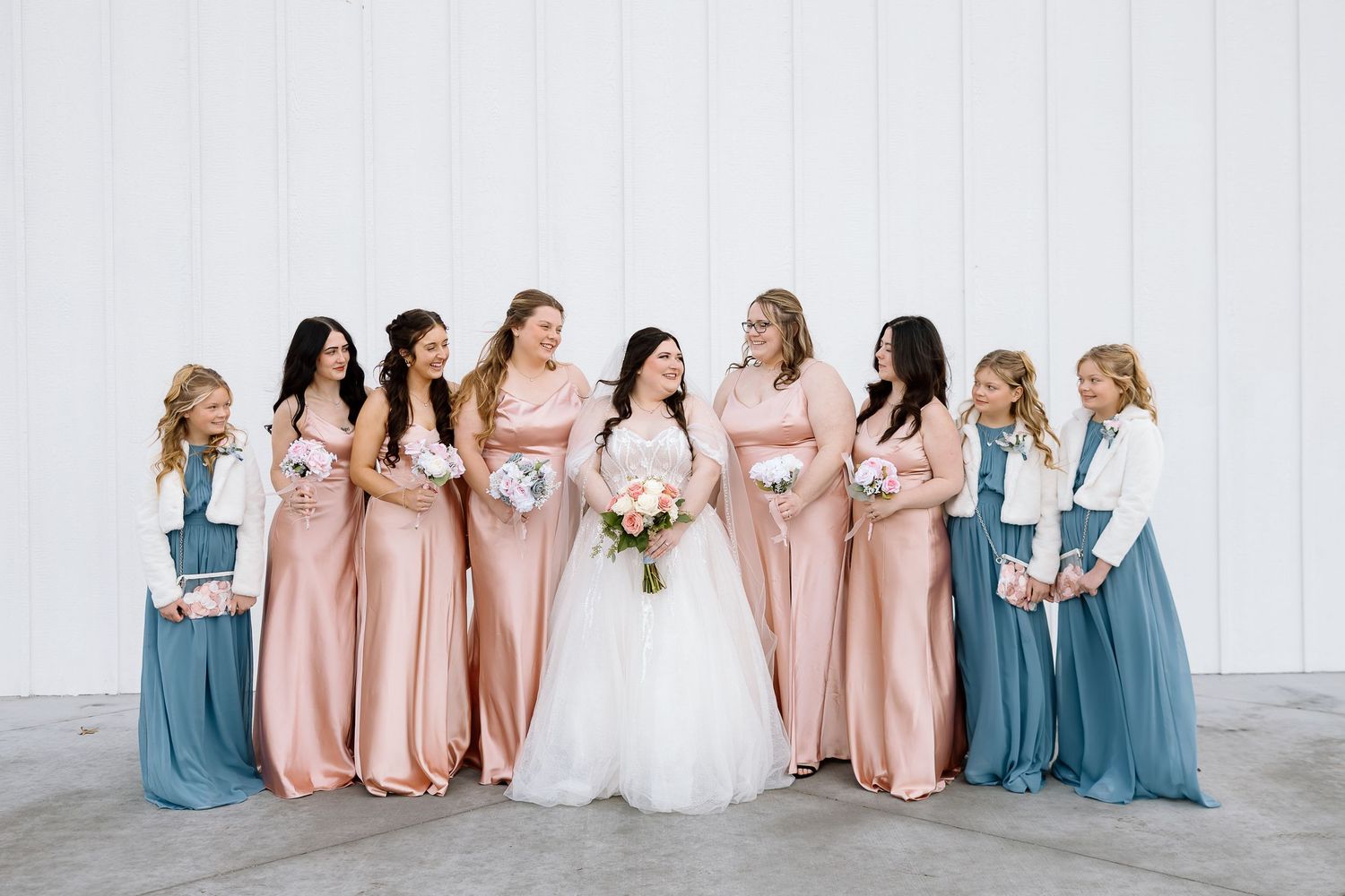 Bridal party poses together wearing elegant blush pink and dusty blue floor-length dresses against white wall.