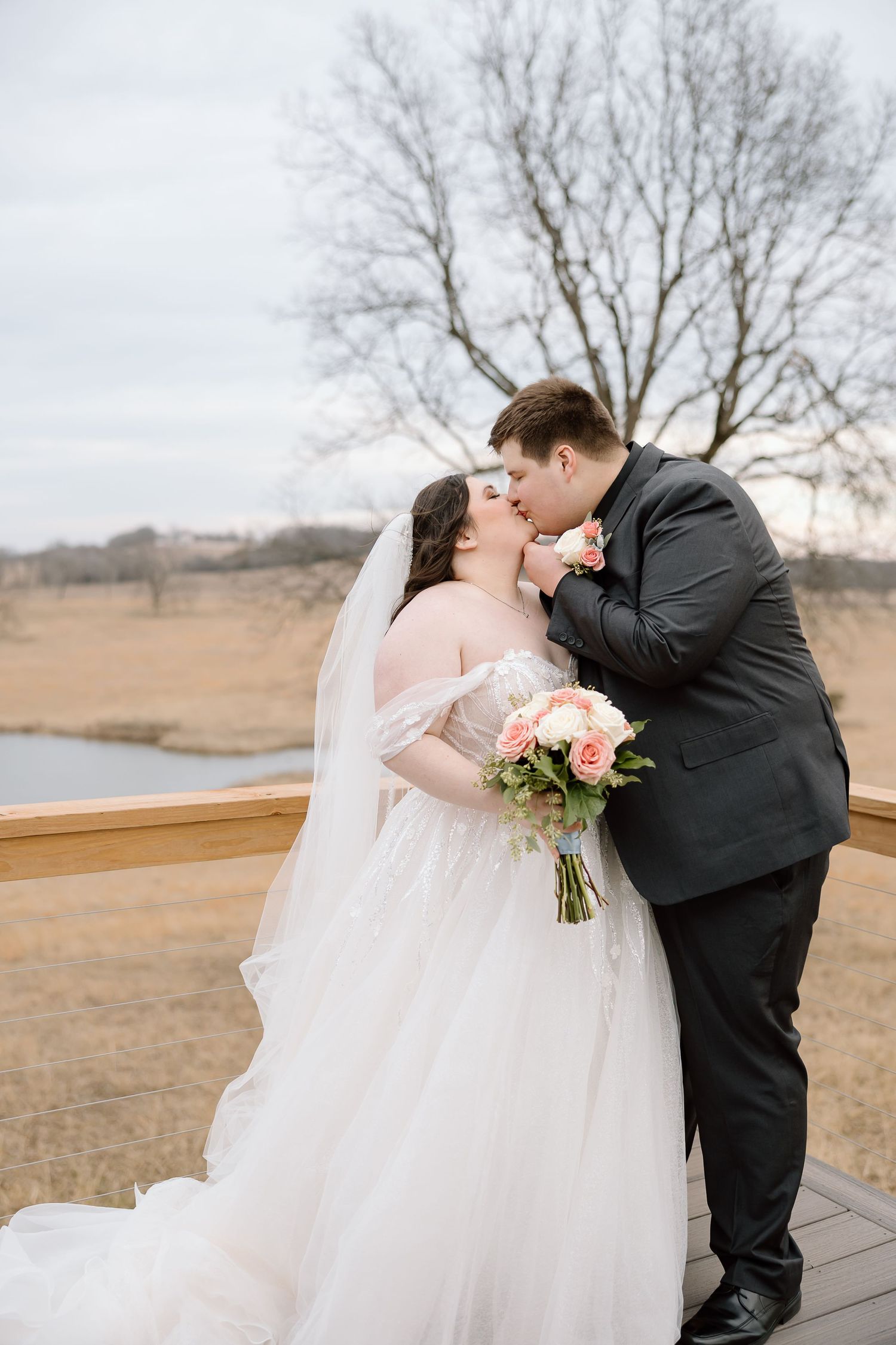 A romantic wedding couple shares a kiss on a wooden deck overlooking a winter landscape with bare trees.