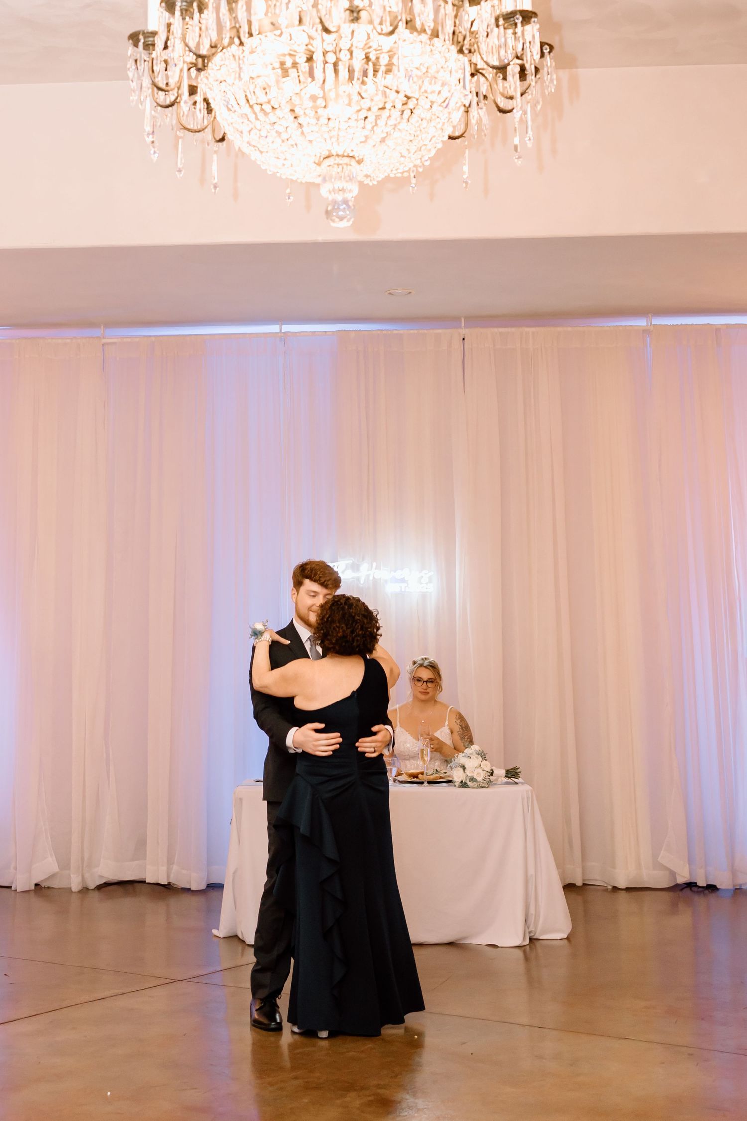 Two people dancing together in a ballroom with white curtains and crystal chandeliers overhead.