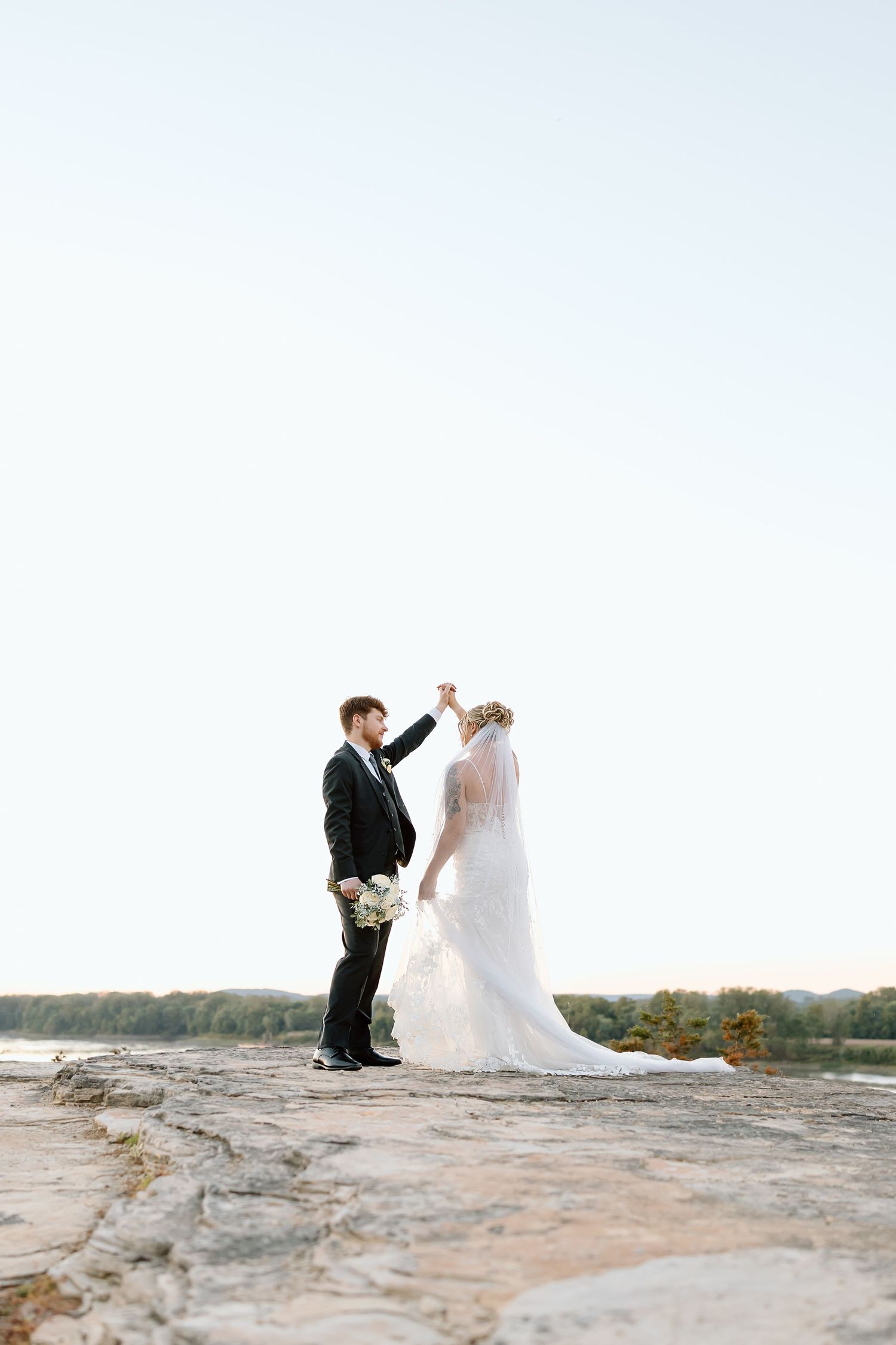 A couple in wedding attire dance together on a scenic rocky overlook during their outdoor celebration.
