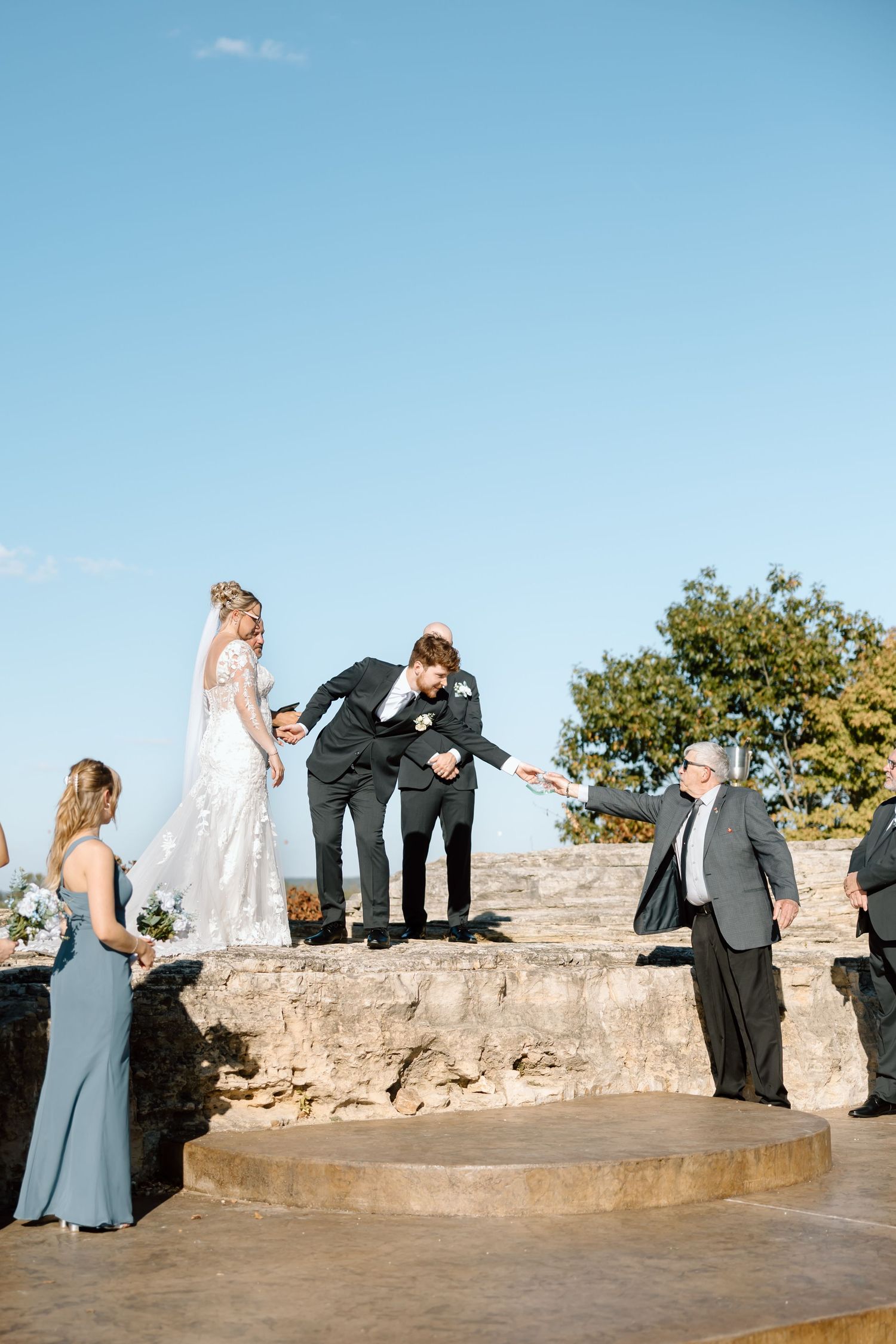 Wedding party members share a candid moment on stone steps against a bright blue sky during an outdoor ceremony.