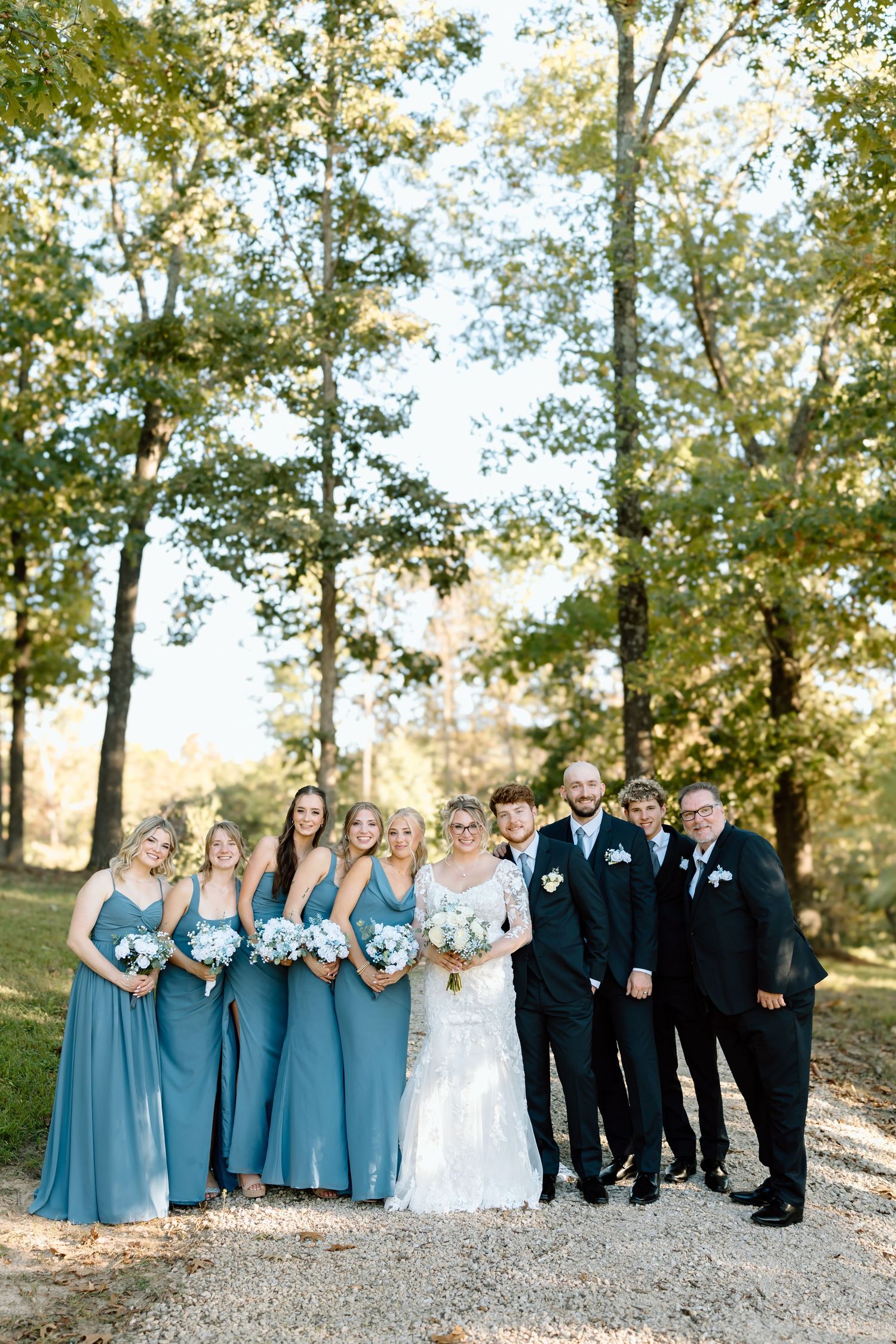 Wedding party in blue bridesmaid dresses and black tuxedos pose together on a gravel path surrounded by autumn trees.