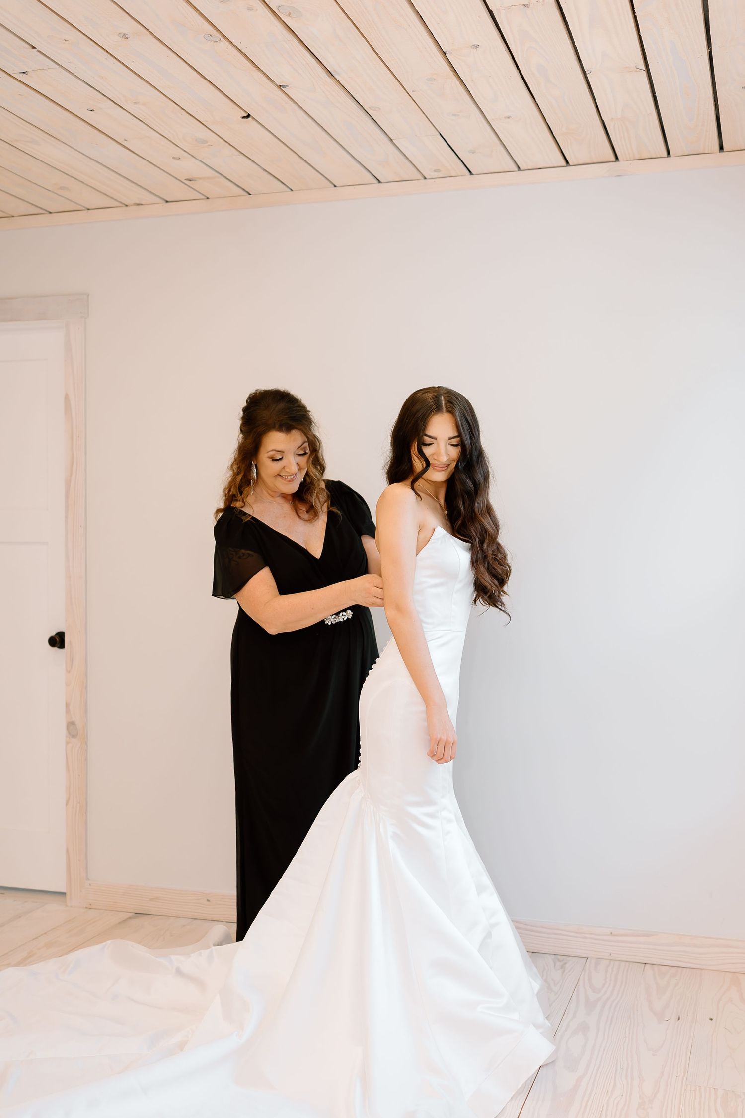 Two women in a minimalist white room, one wearing a black dress helping adjust a white mermaid-style wedding gown.