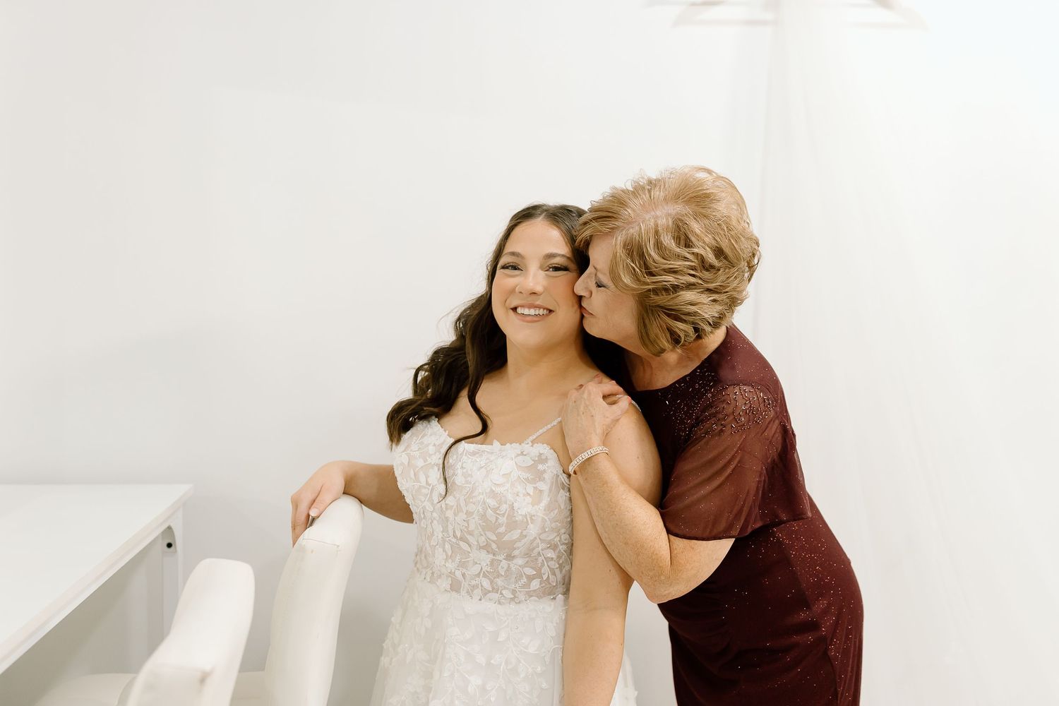 Bride in white wedding dress embraces loved one in burgundy dress during emotional wedding moment.