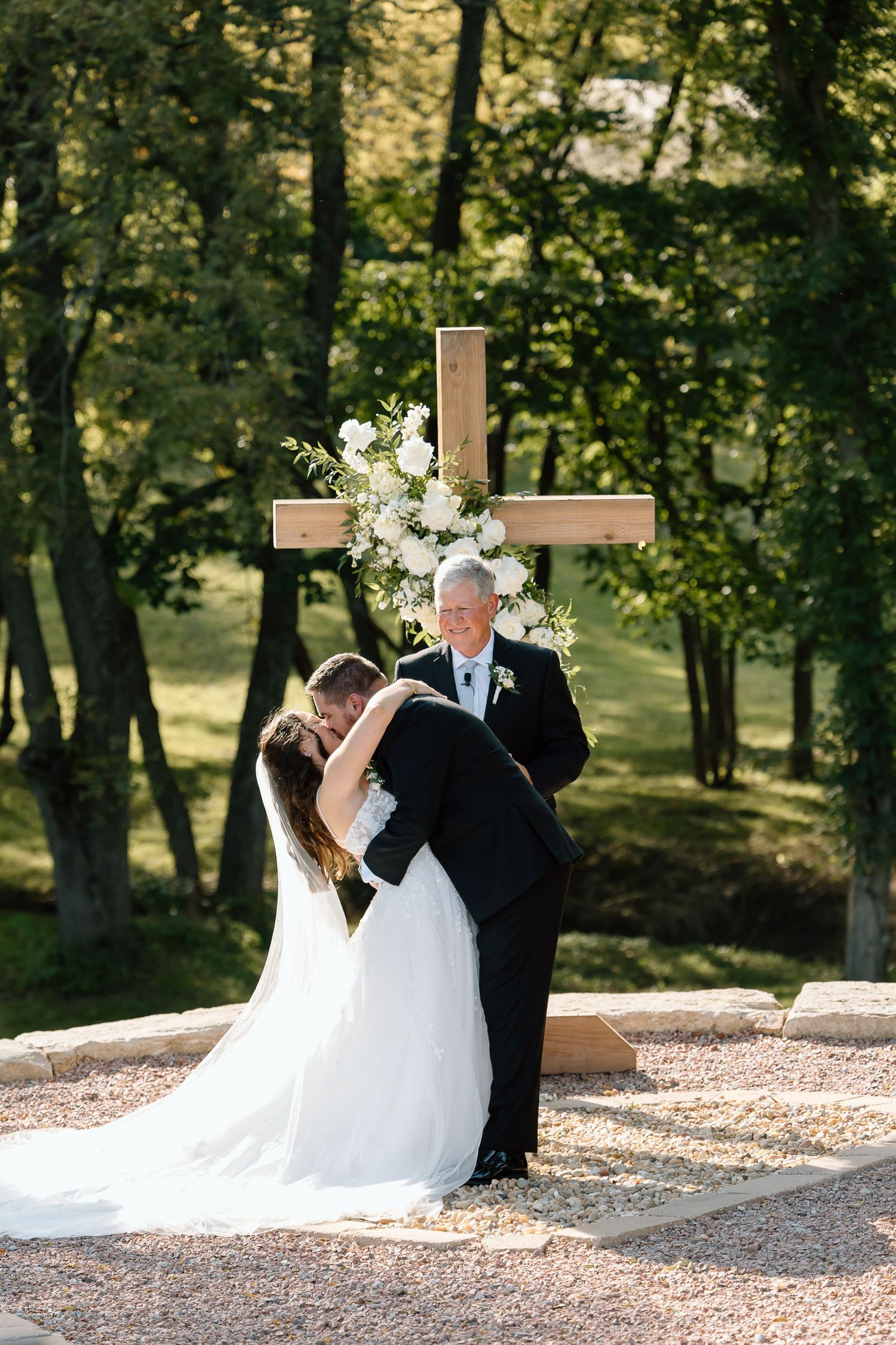 A wedding ceremony takes place outdoors with a wooden cross adorned with white flowers in a serene wooded setting.