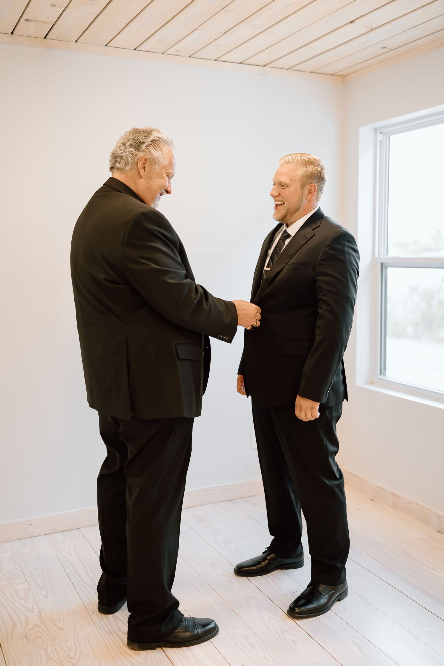 Two people in black suits stand in an empty white room with wood floors having a conversation.