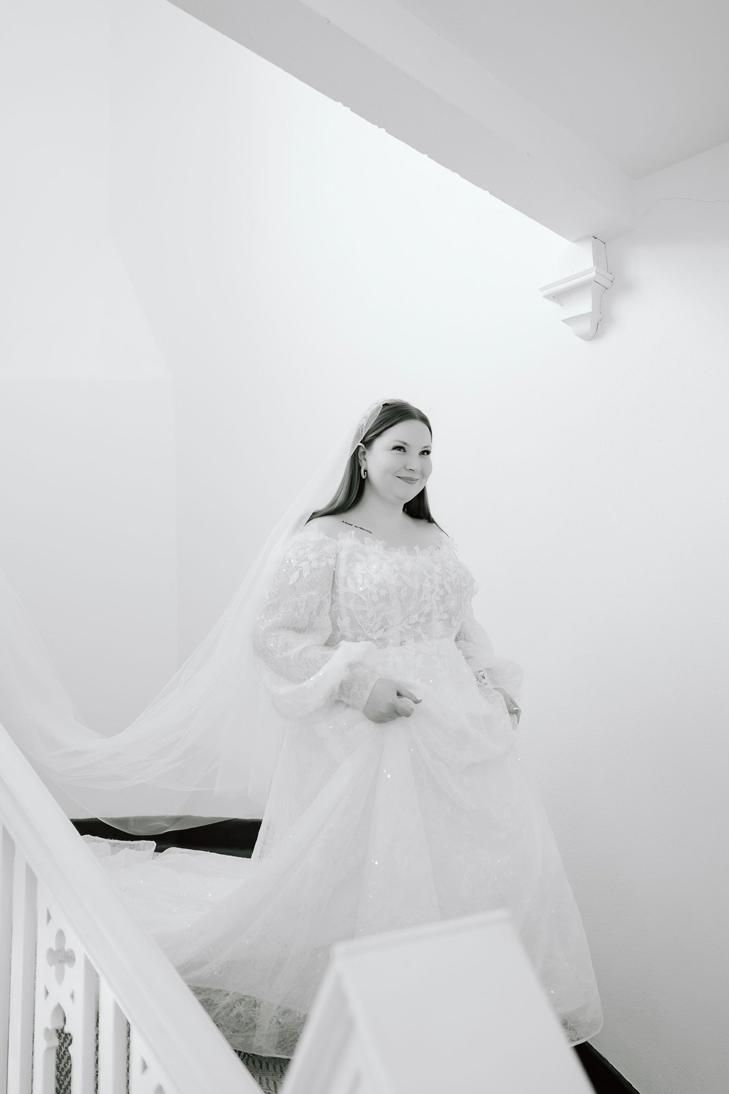Bride in white lace gown descending staircase, black and white wedding photo.