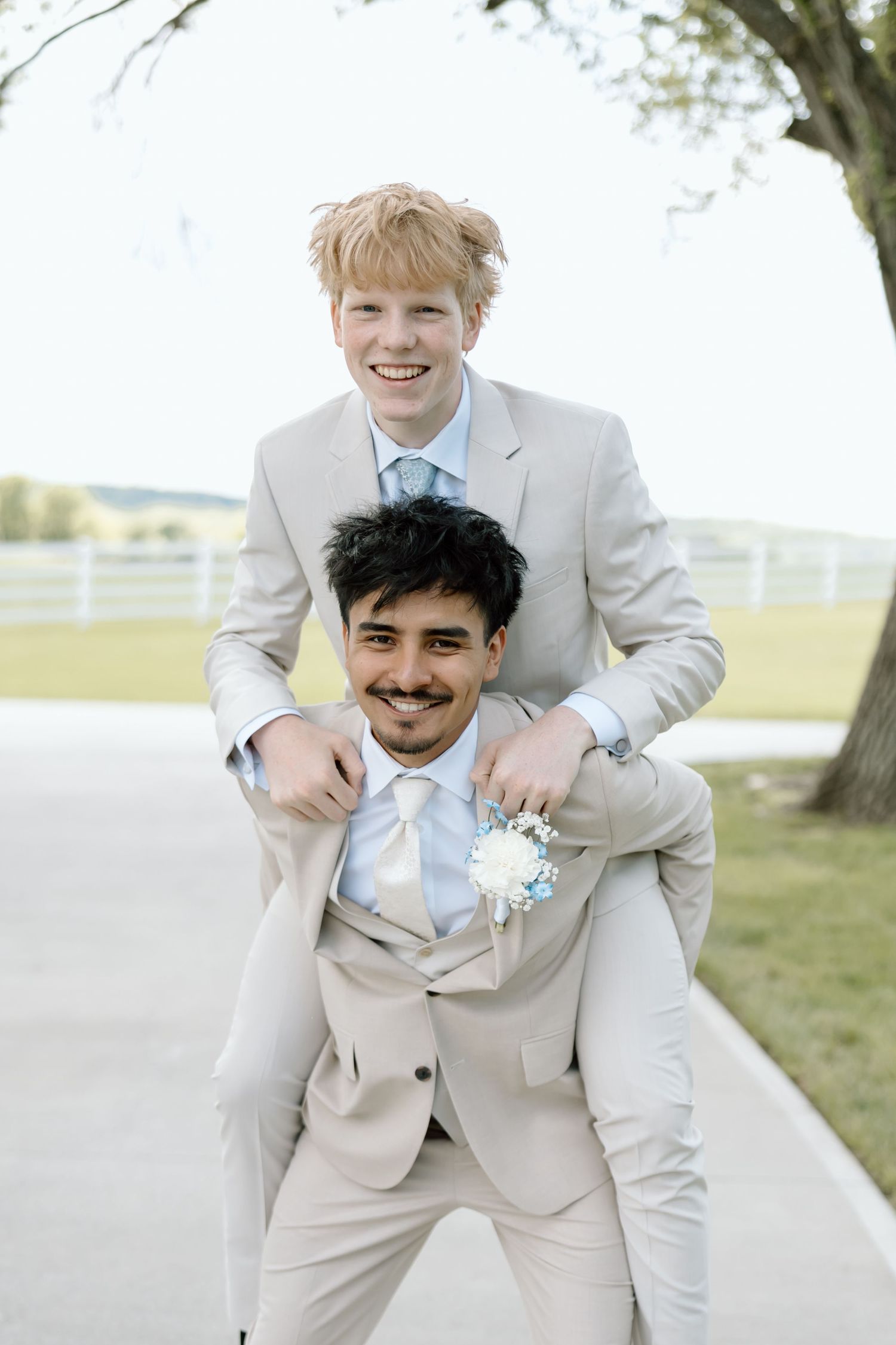 Two young men in matching beige suits smile outdoors, one giving the other a piggyback ride at a formal event.