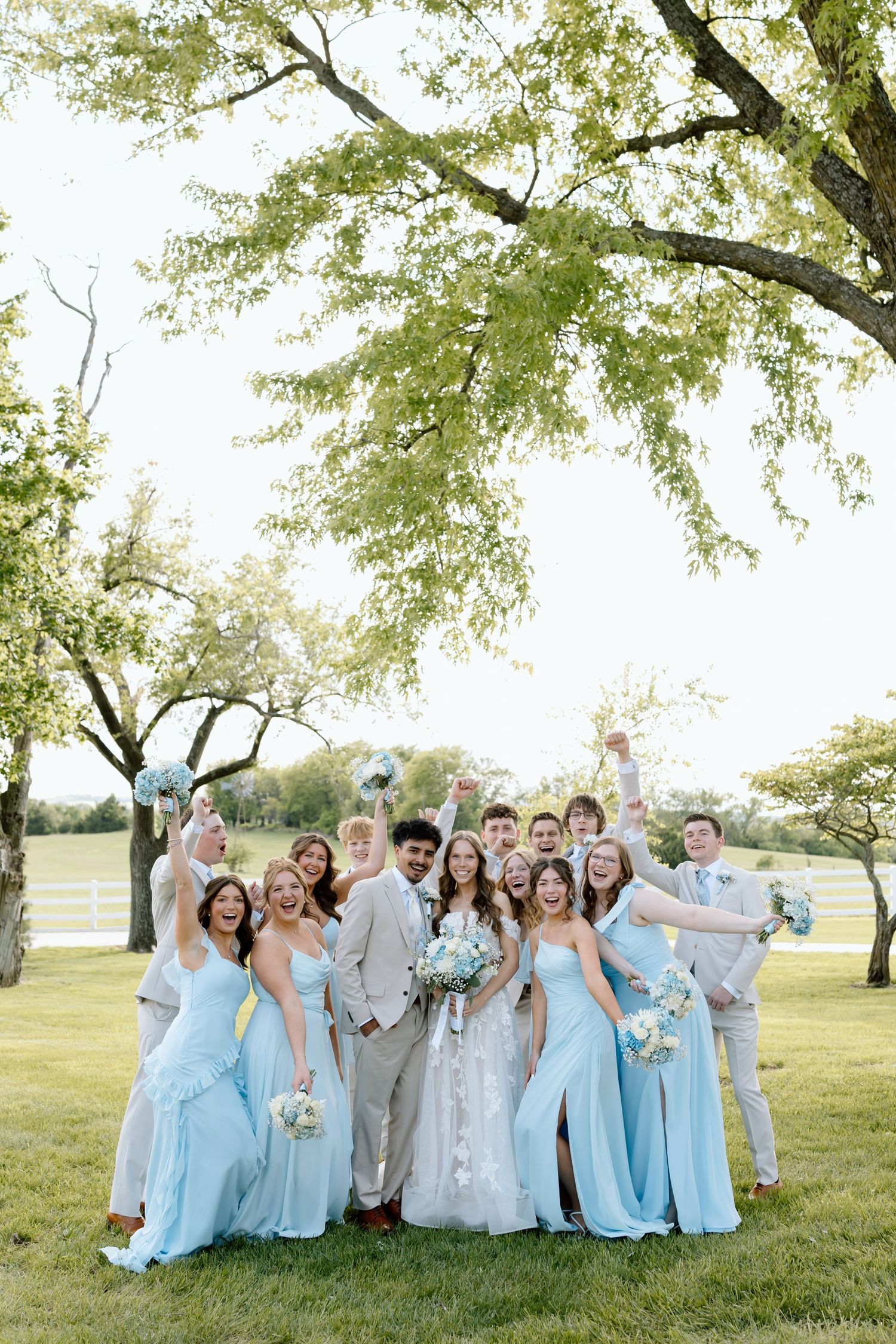 Wedding party celebrating outdoors in light blue bridesmaid dresses and tan suits under green trees on a sunny day.