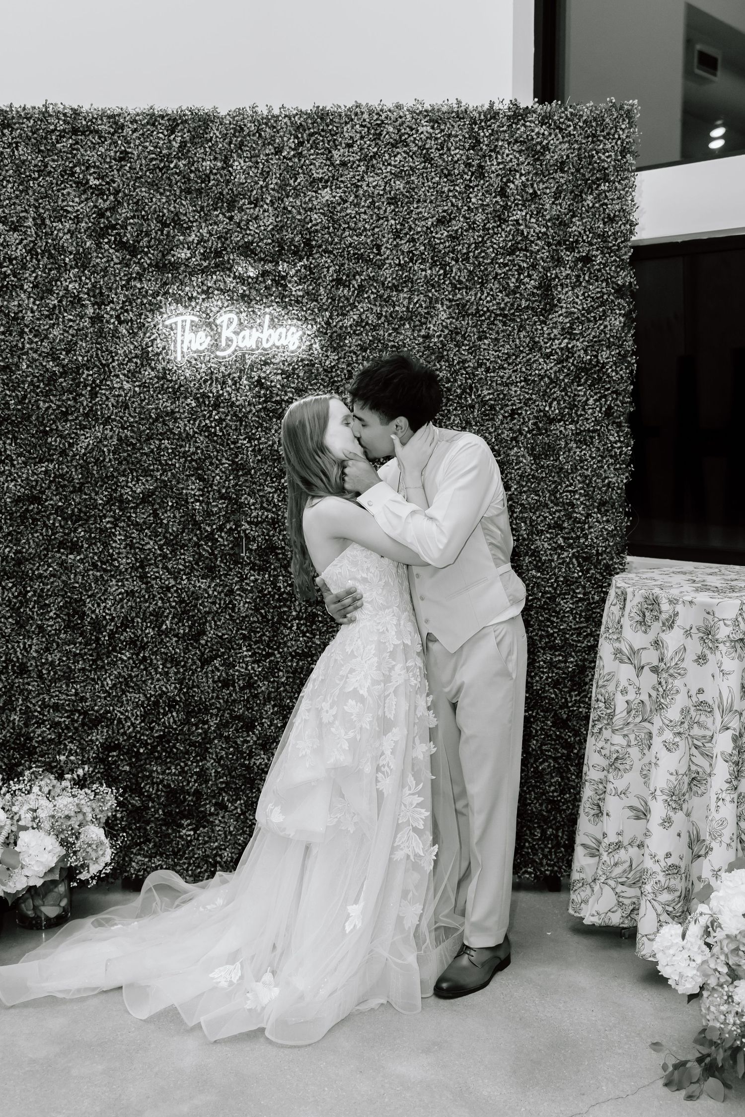 Black and white photo of a bride and groom sharing a kiss in front of a lush greenery wall with a neon sign.