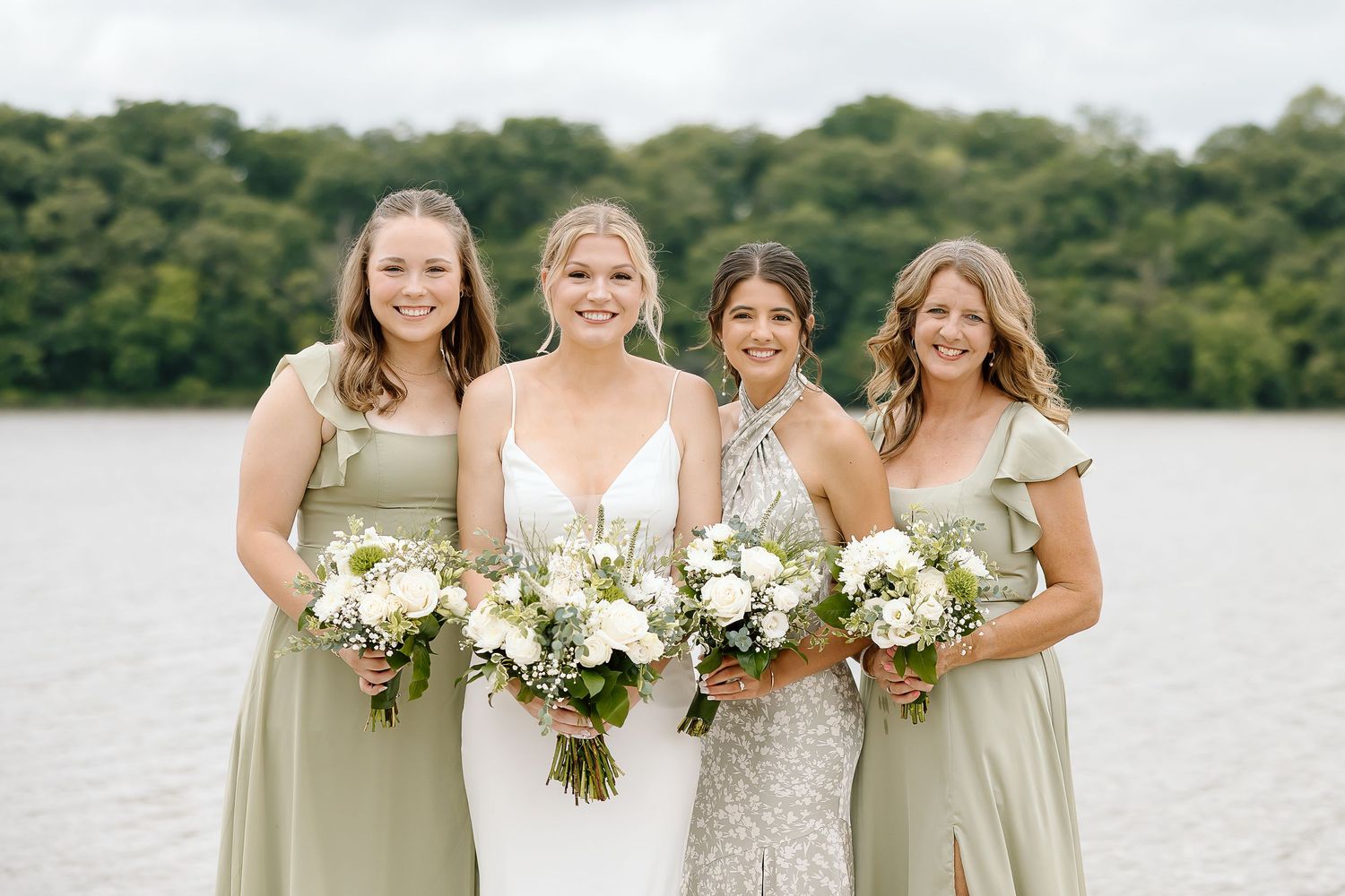 Four women in sage green and white dresses hold elegant white floral bouquets while posing by a serene lakeside.