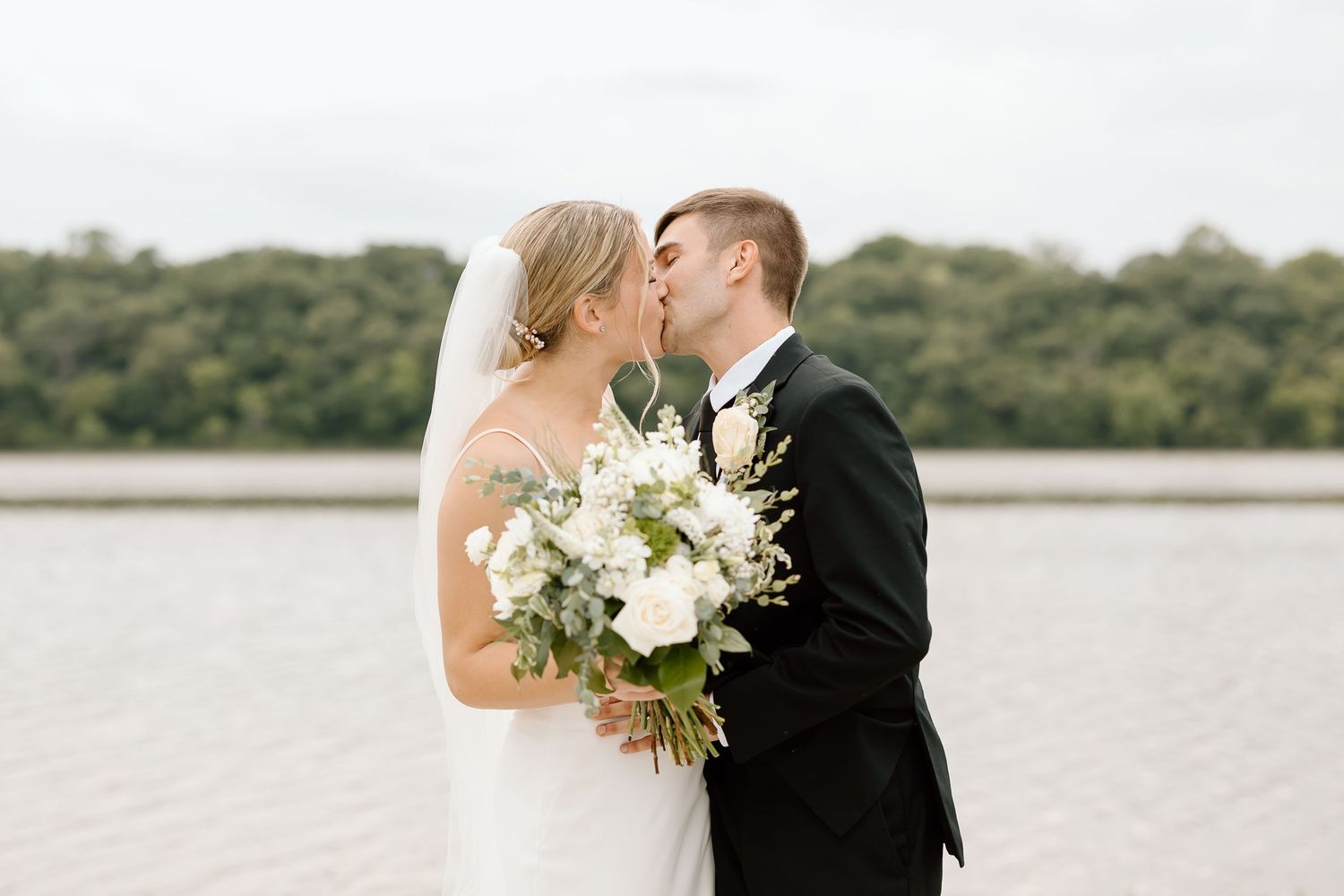 A romantic wedding couple kisses while holding an elegant white floral bouquet by a lakeside on their special day.