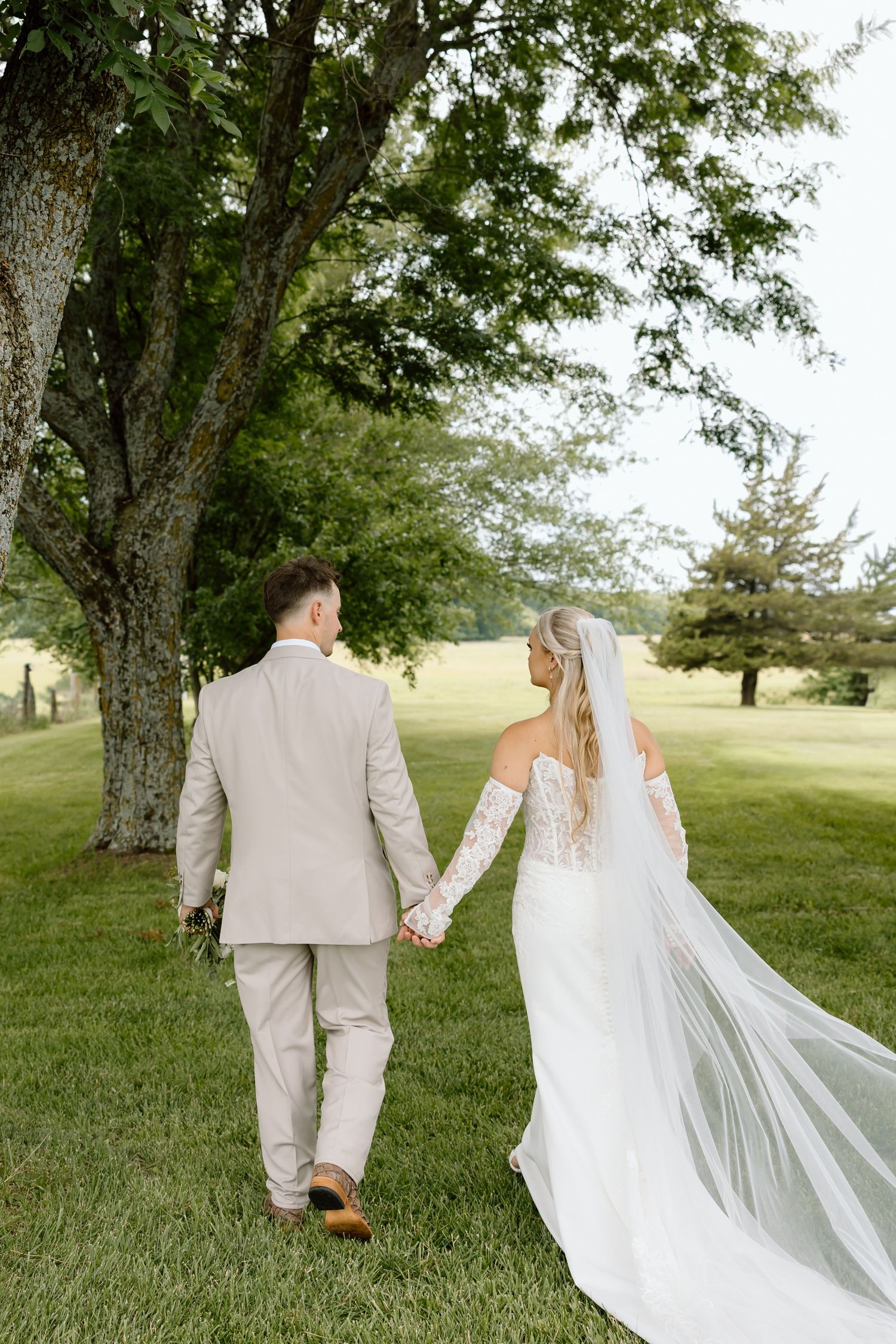 Bride and groom walking hand in hand through lush green garden, bride wearing lace gown with flowing veil.