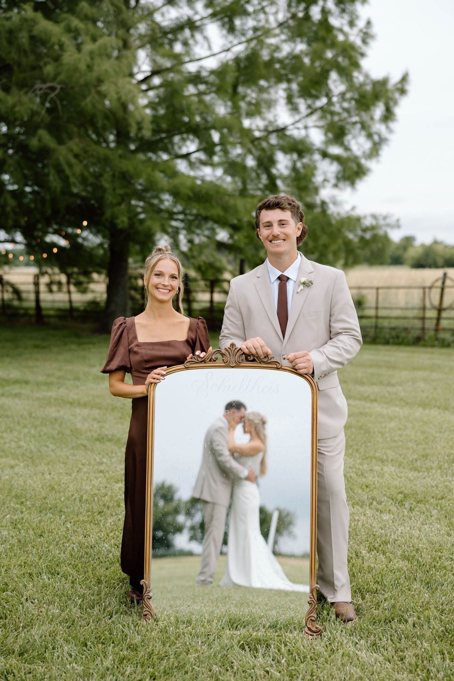 Couple holding ornate gold mirror reflecting kissing bride and groom at outdoor wedding venue.