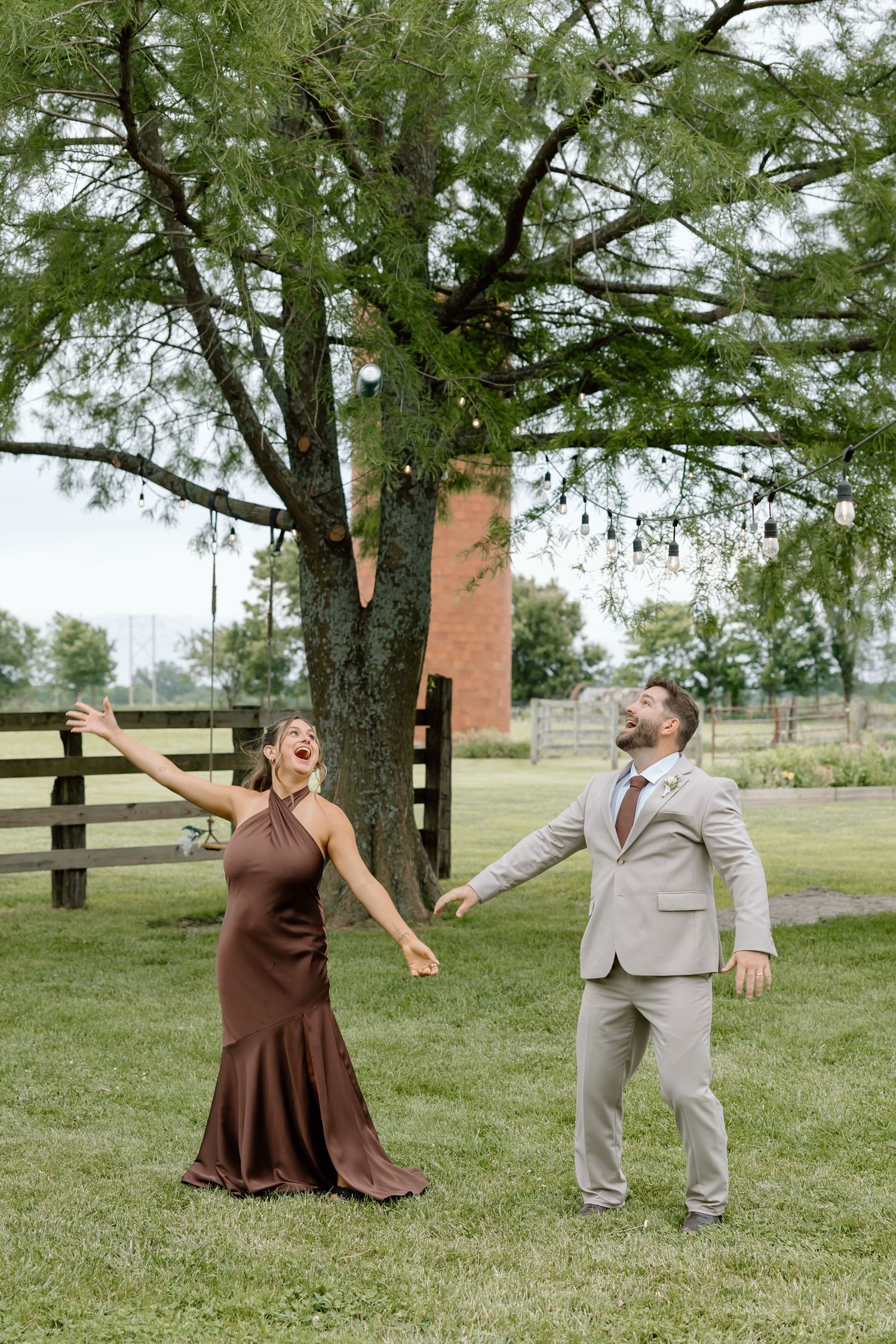 A joyful couple in formal attire laughing outdoors near a large tree on a green lawn at a wedding venue.