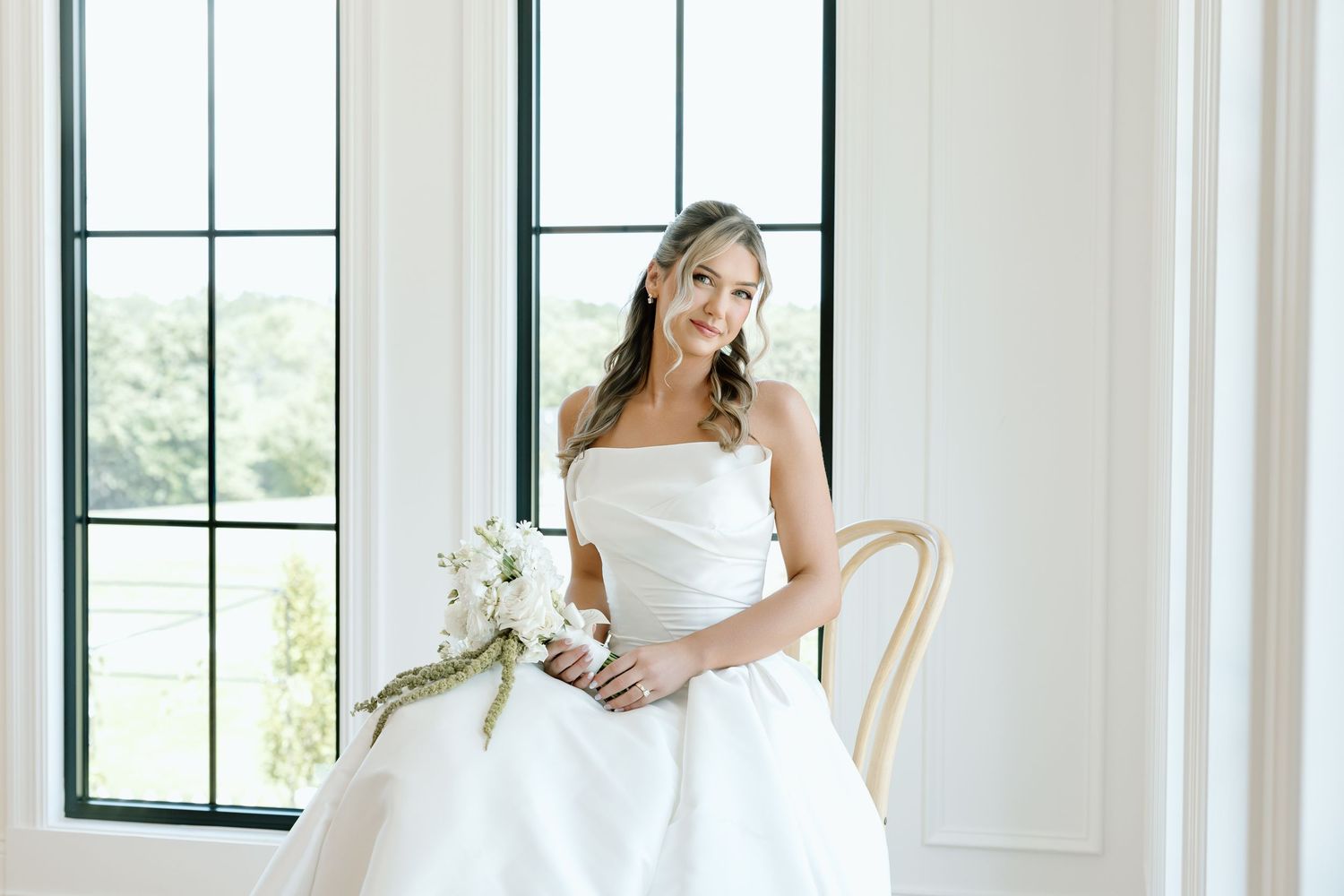 Bride in strapless white ball gown holding white bouquet, seated by large windows in bright elegant room.