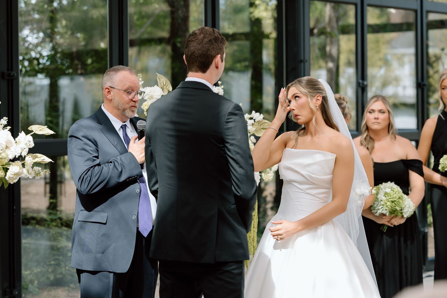 Emotional bride wiping tears during outdoor wedding ceremony with groom and officiant, bridesmaid visible in background.