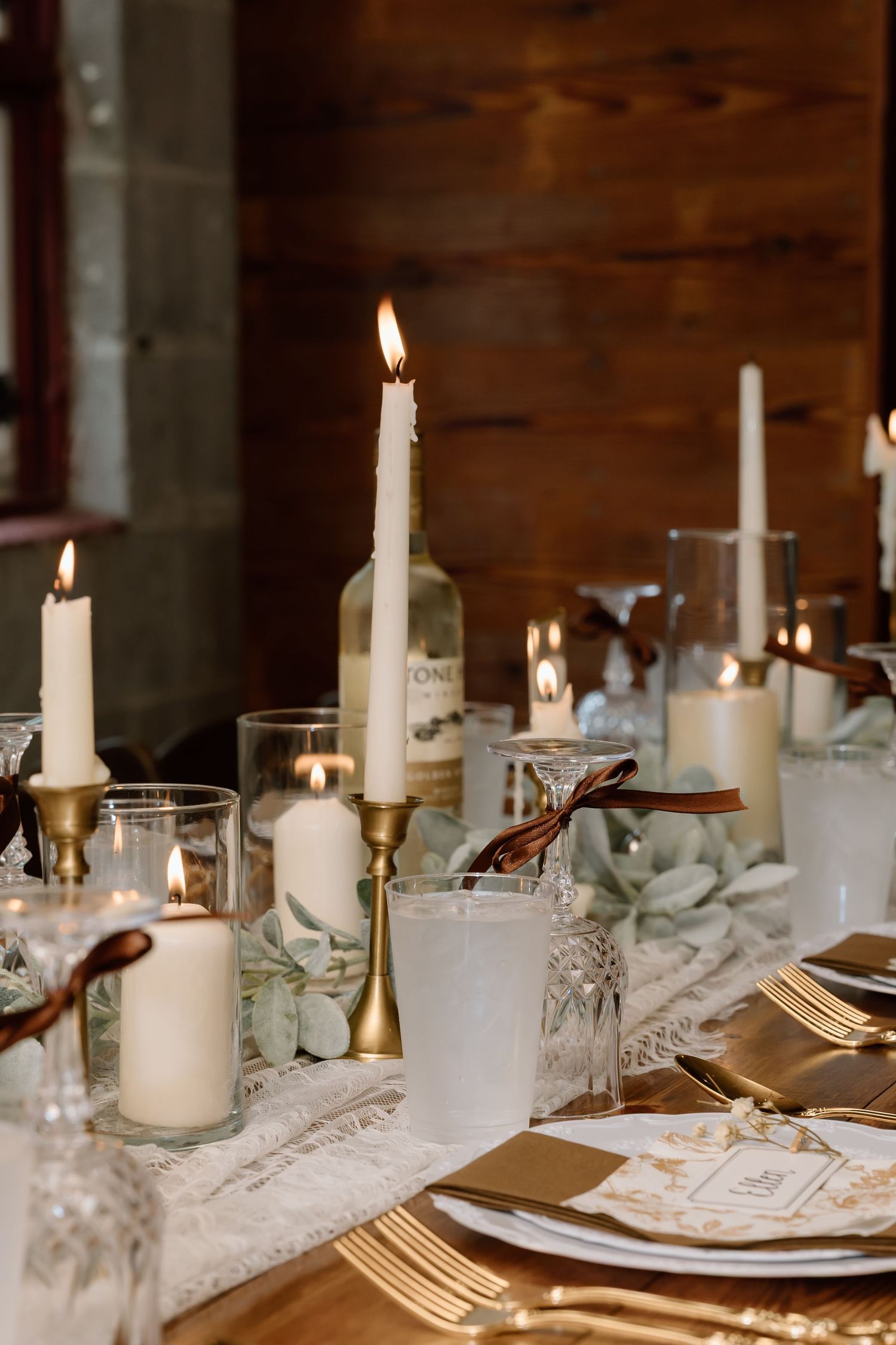 Elegant wedding table setting with gold candlesticks, white taper candles, crystal glasses, and lace runner in rustic barn.