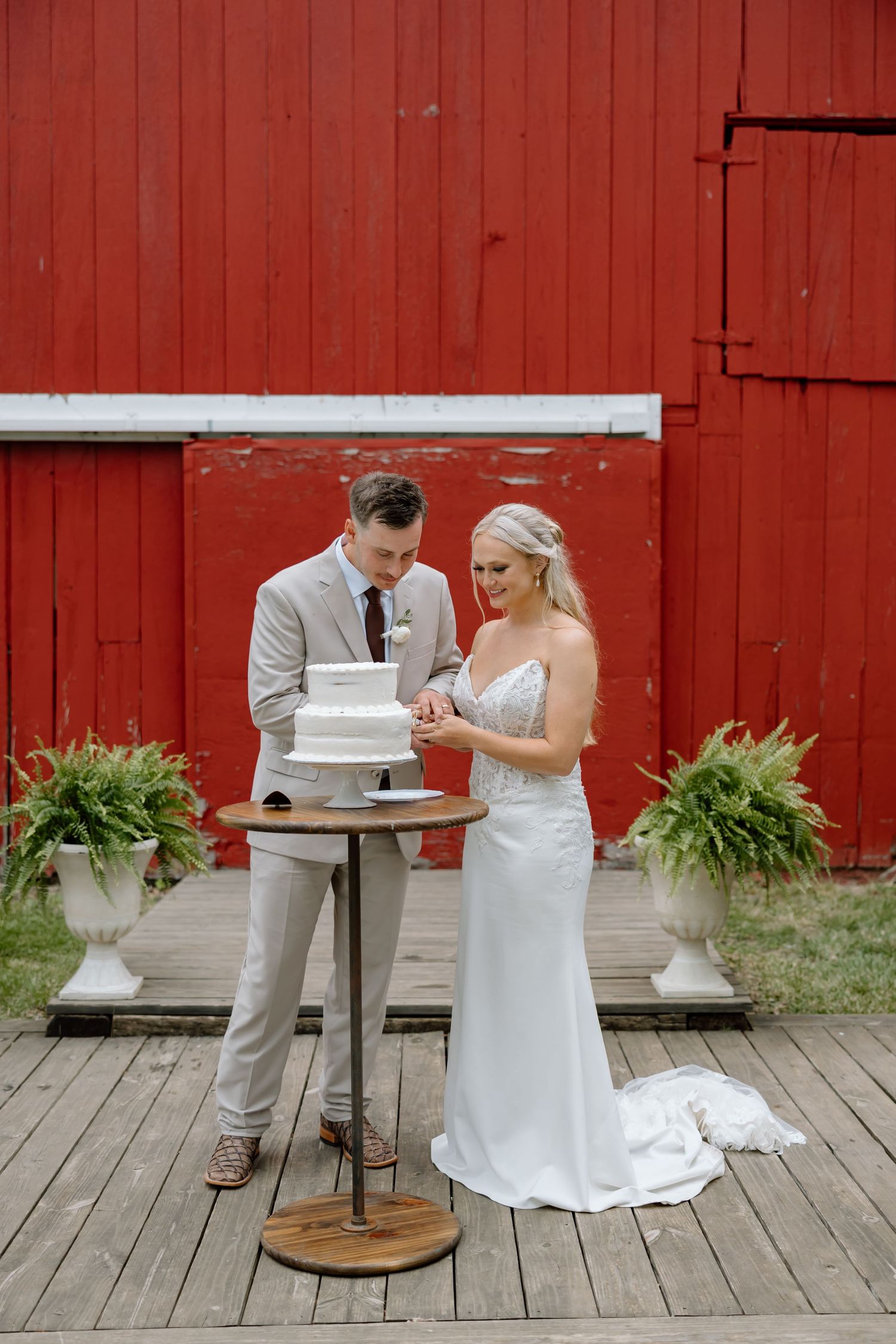 Bride and groom cutting white wedding cake on wooden deck in front of red barn with fern plants.