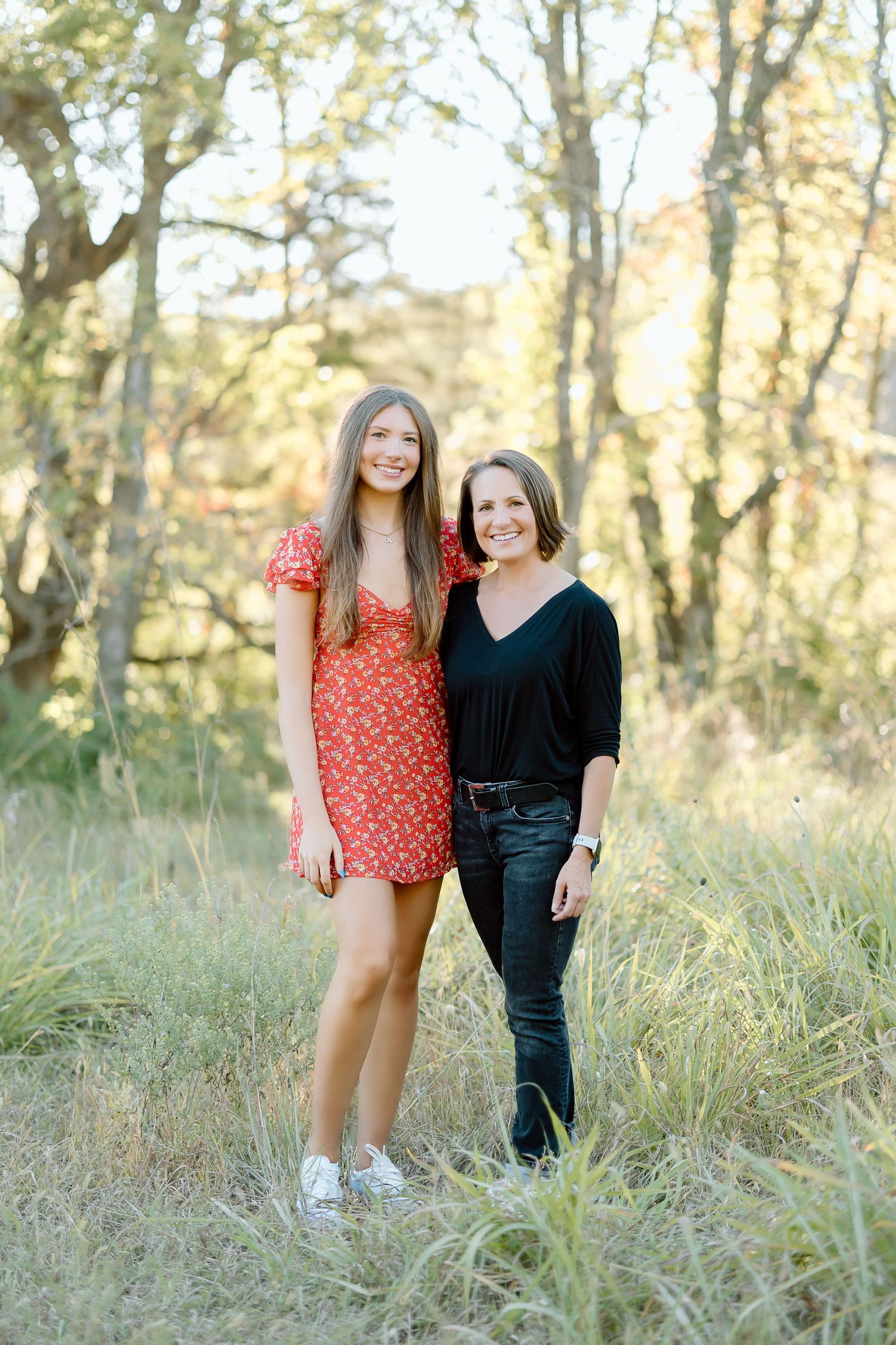 Two friends in contrasting outfits stand together in a sunlit field with tall grass and trees in the background.
