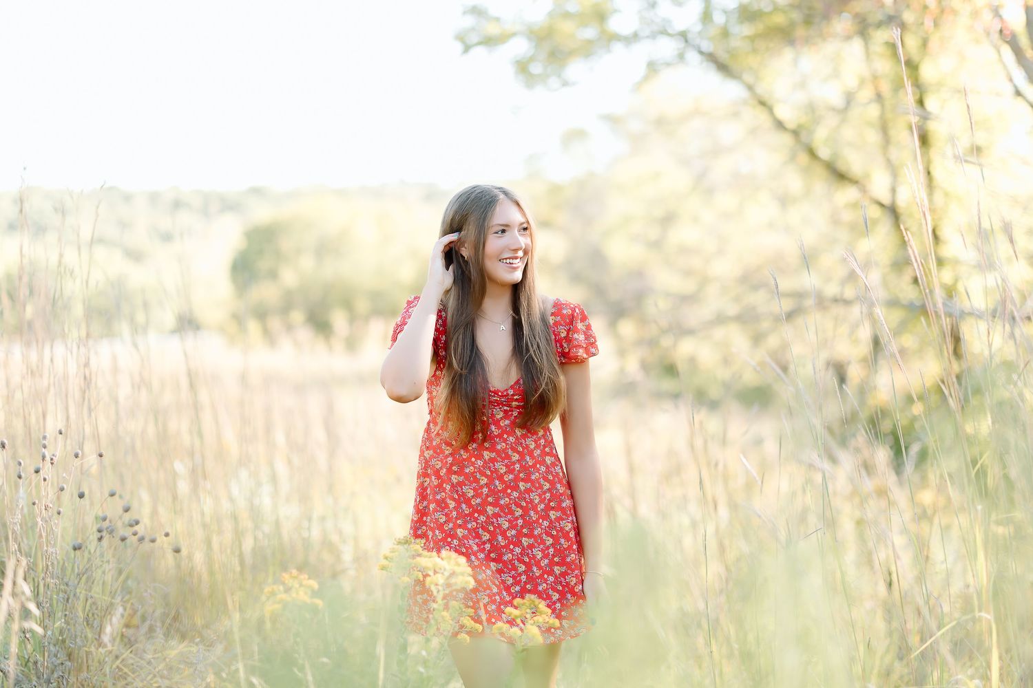 A person in a red floral dress walks through a sun-lit field of tall golden grass at sunset.