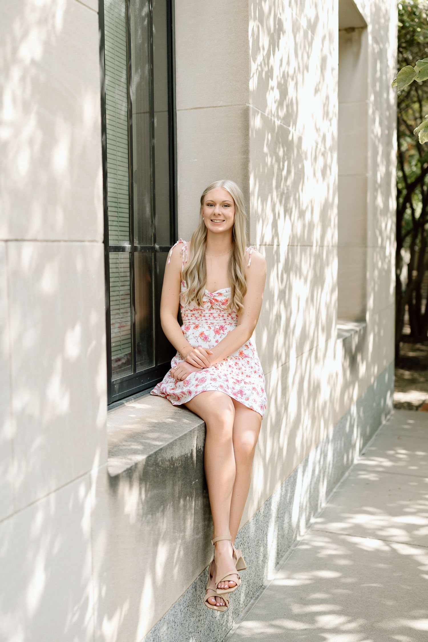 A person in a floral dress poses against a white wall with green shutters in dappled sunlight.