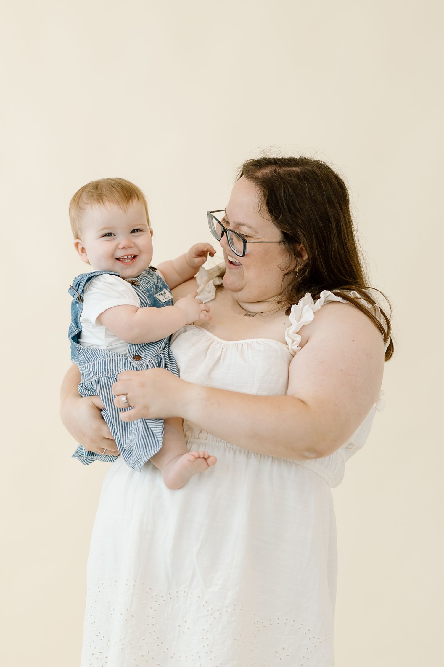 A woman in a white dress holds a baby wearing blue overalls against a neutral beige background.