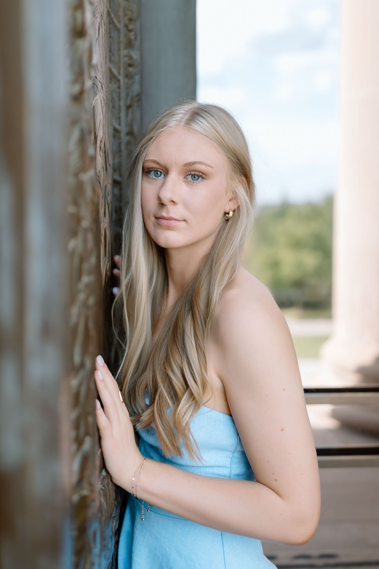 A woman in a light blue strapless dress poses against a pillar outdoors with soft natural lighting.