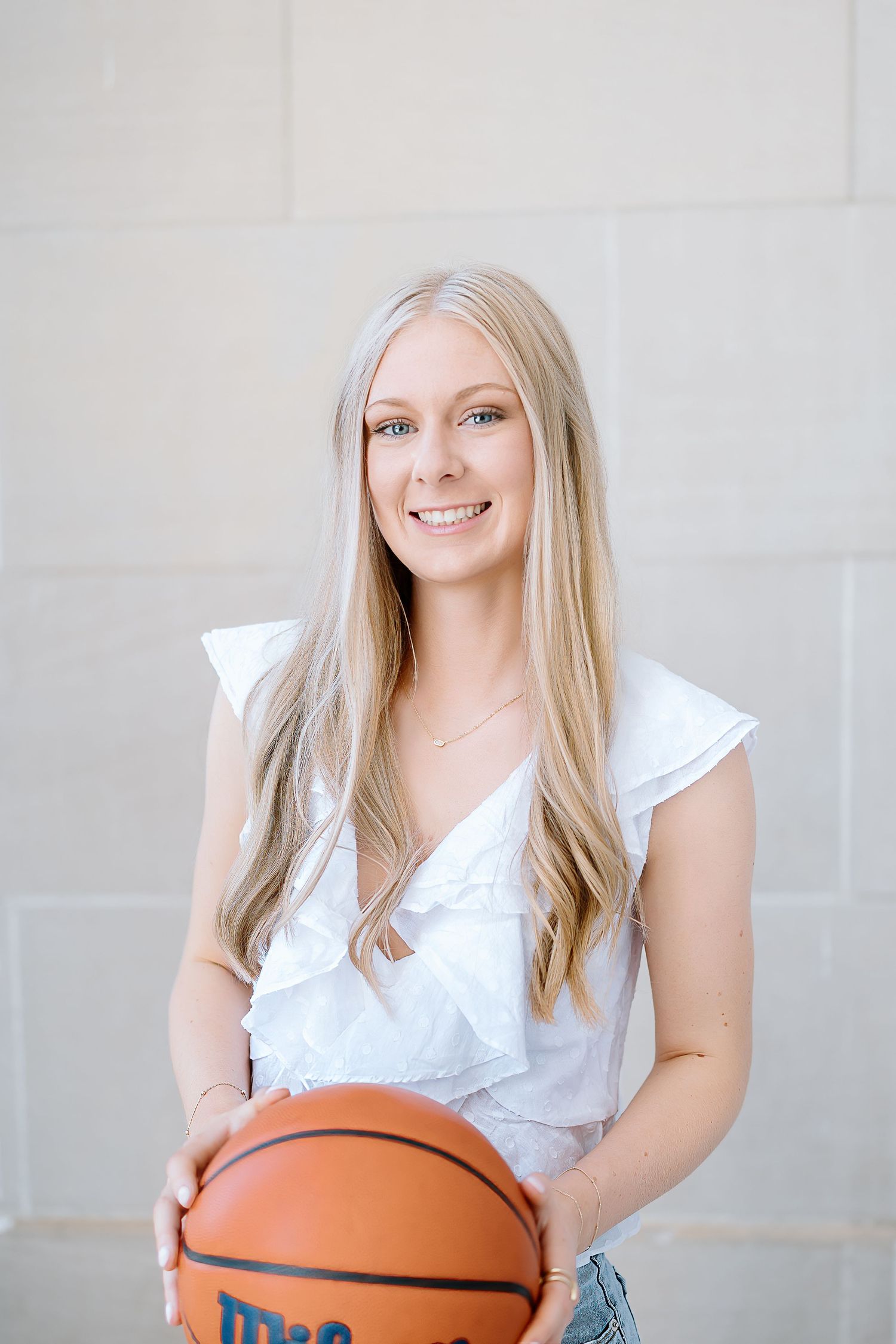 Basketball player in white ruffled top poses with orange basketball against light gray background.