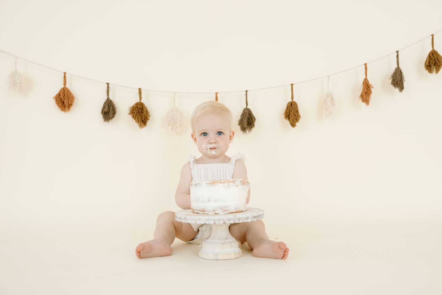 A baby in a white dress sits on a light backdrop with hanging decorative ornaments above.