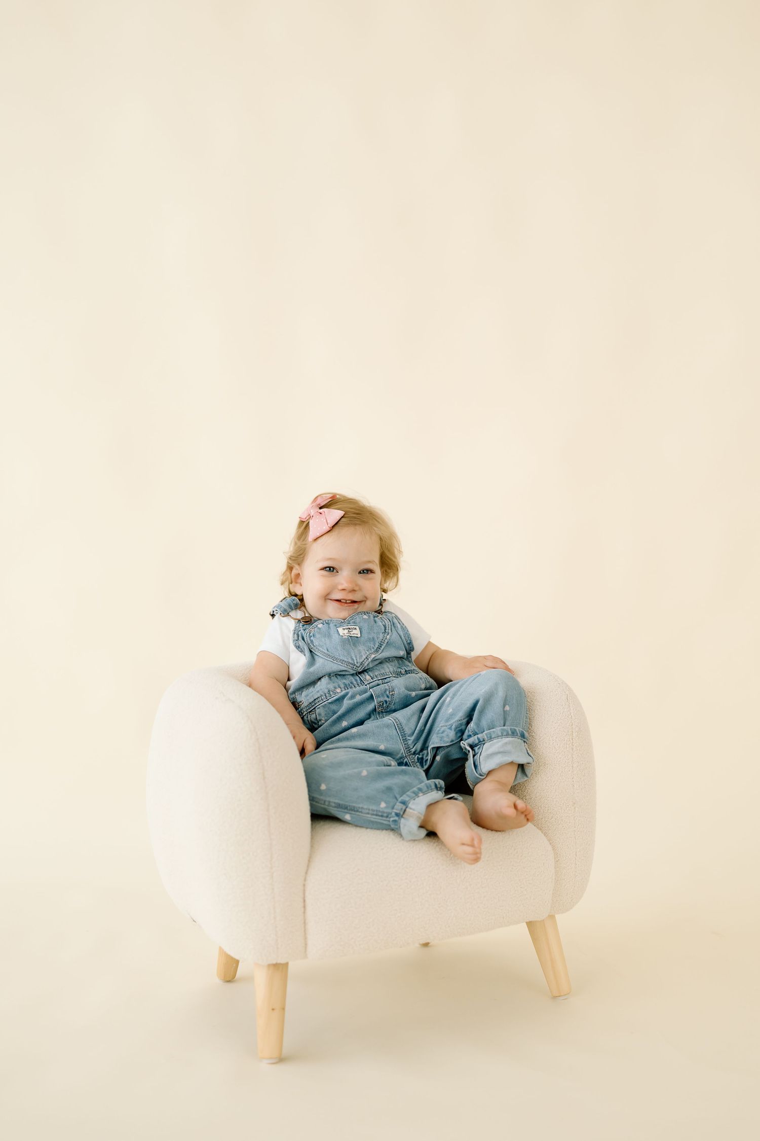A small child in denim overalls sits and plays on a modern white curved chair with wooden legs.