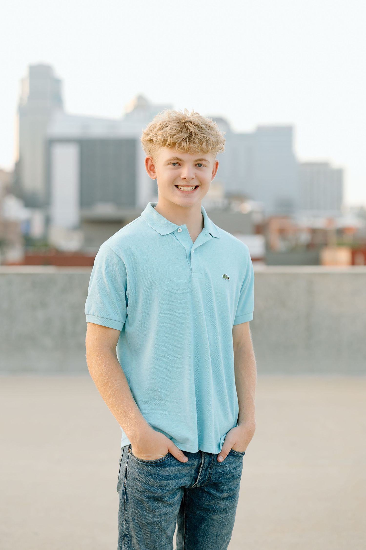 A person in a light blue polo shirt and jeans poses casually against an urban cityscape background.