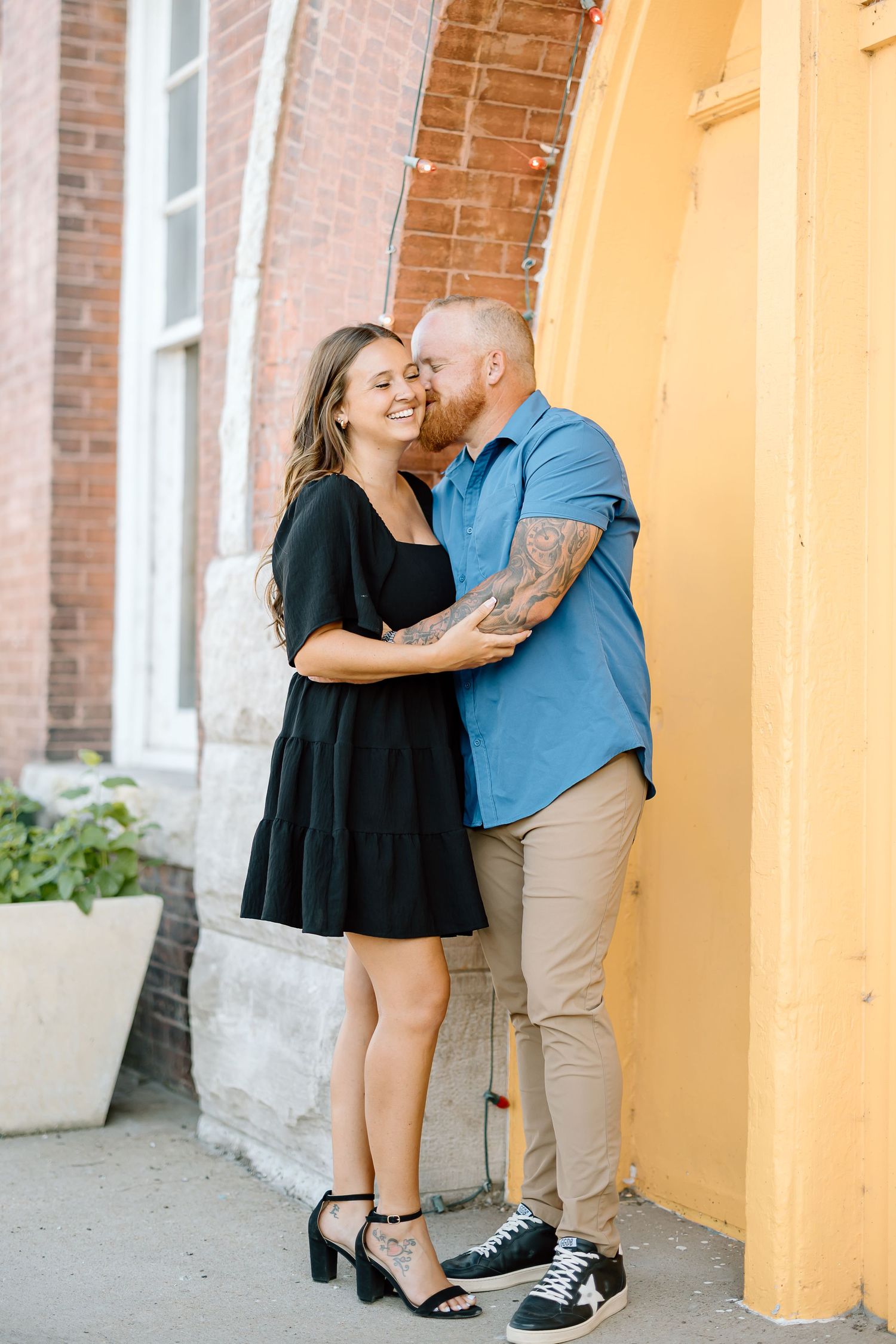 A couple shares an intimate moment against a bright orange wall with brick details in an urban setting.