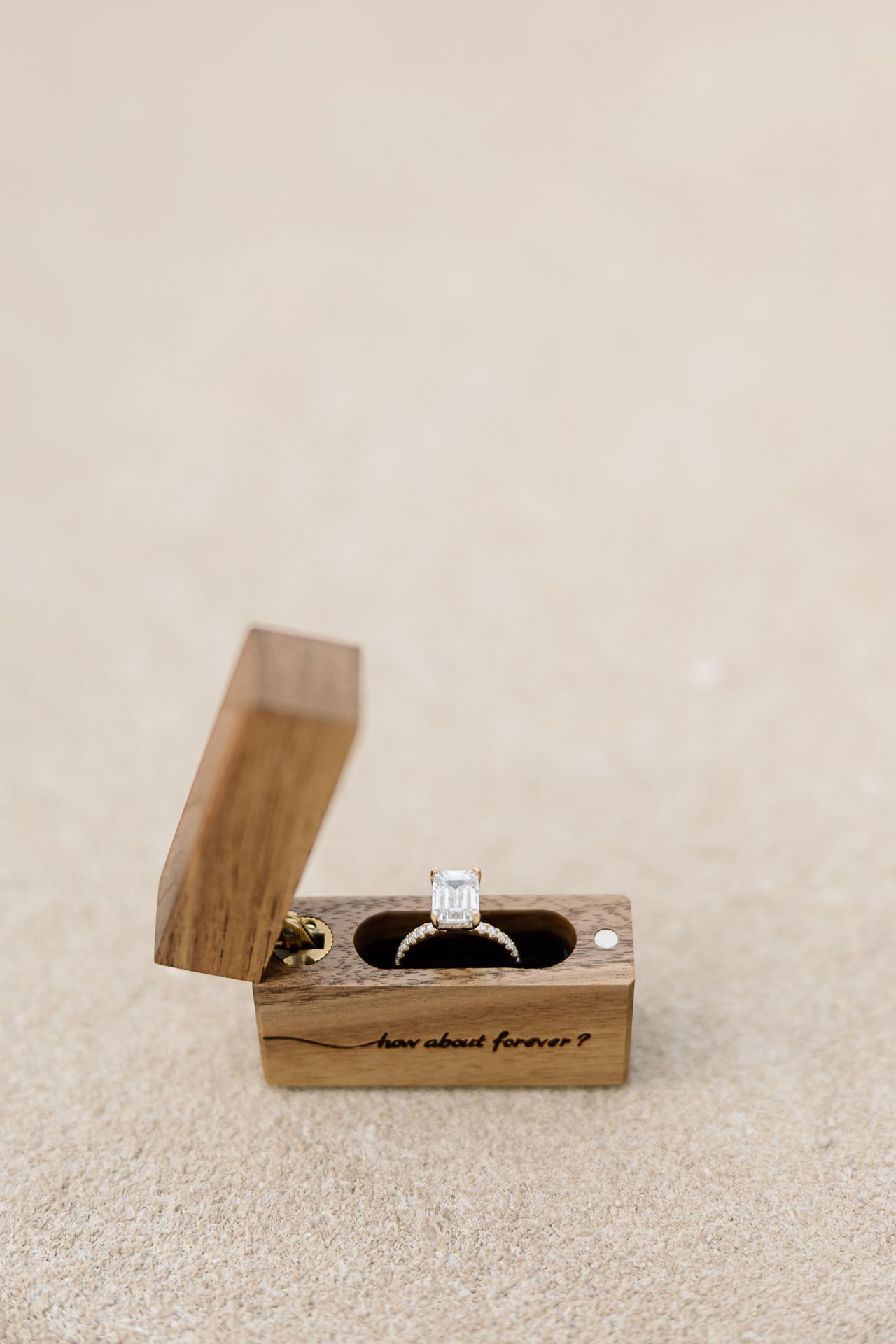 Diamond engagement ring displayed in small wooden box with hinged lid on beige textured surface.