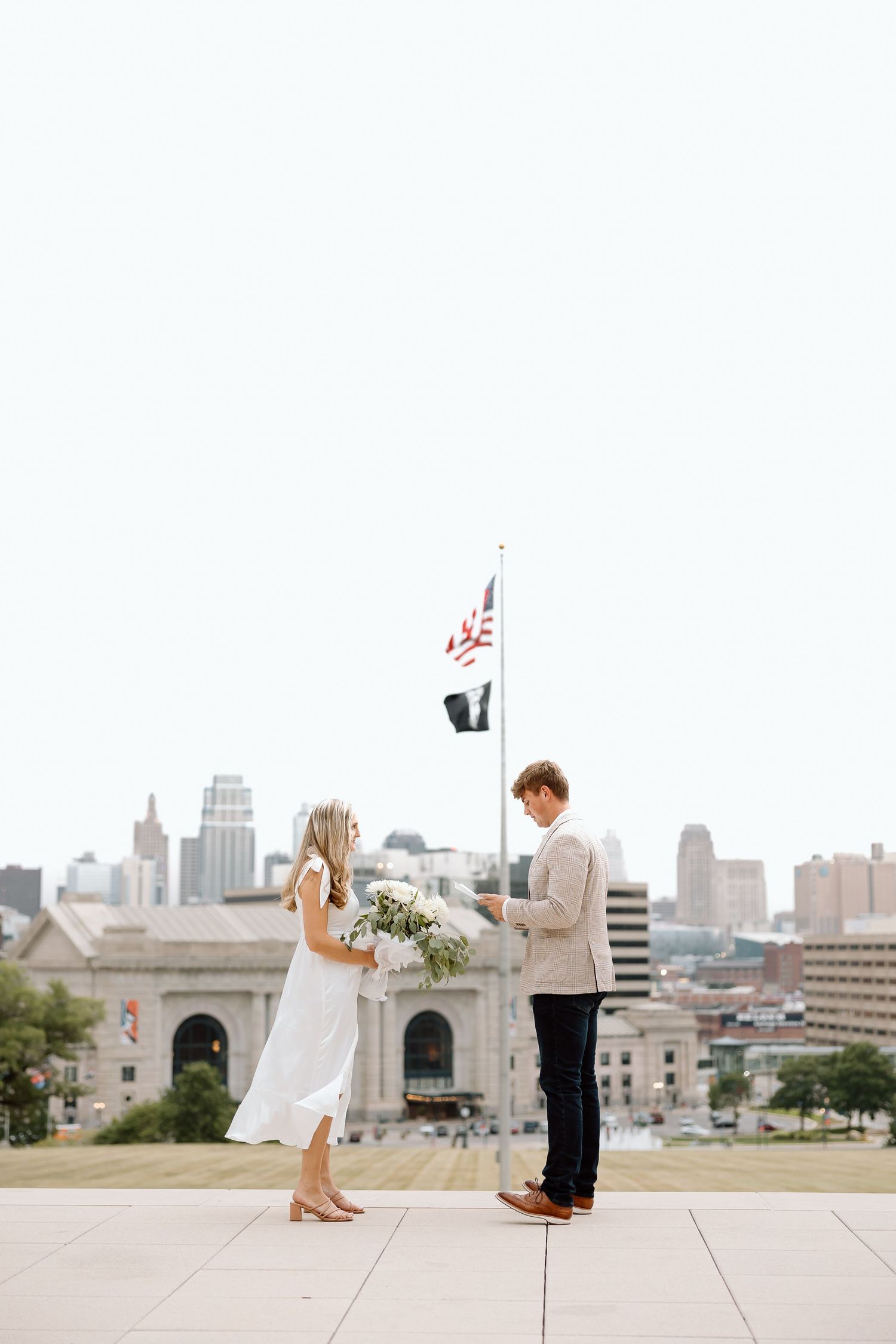 A couple stands on a terrace overlooking the Kansas City skyline during a romantic moment with urban architecture behind them.