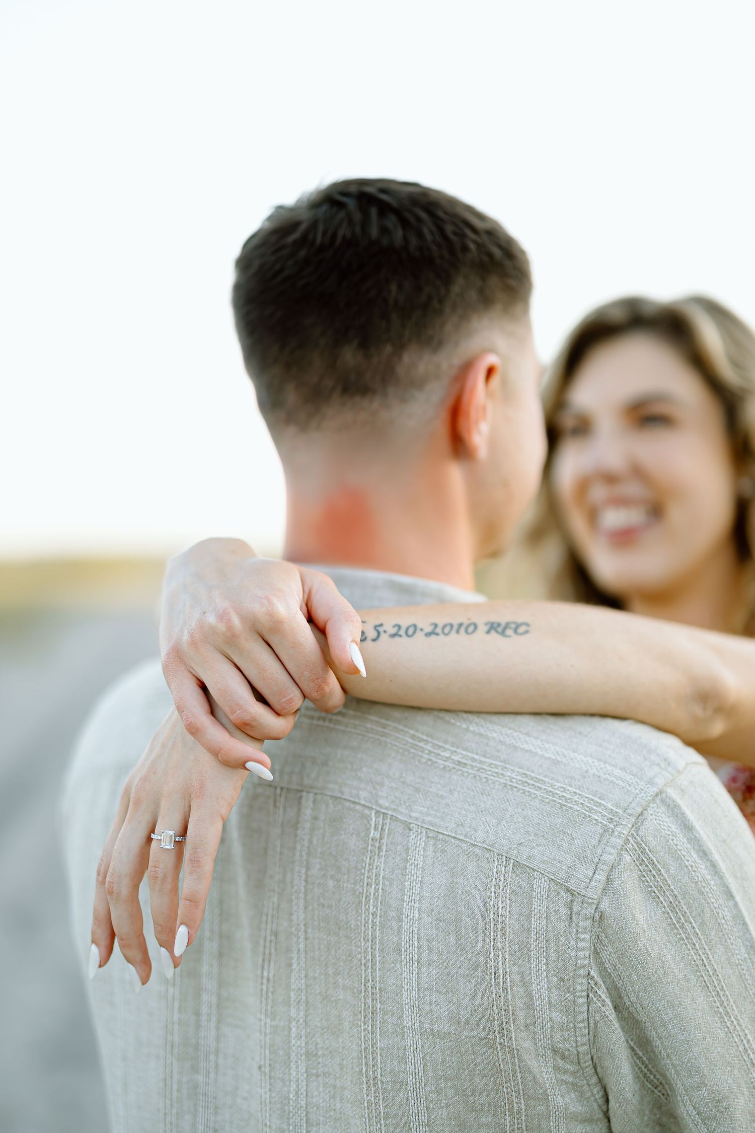 Close-up shot of an engagement ring on a hand with a soft blurred background showing a couple embracing.