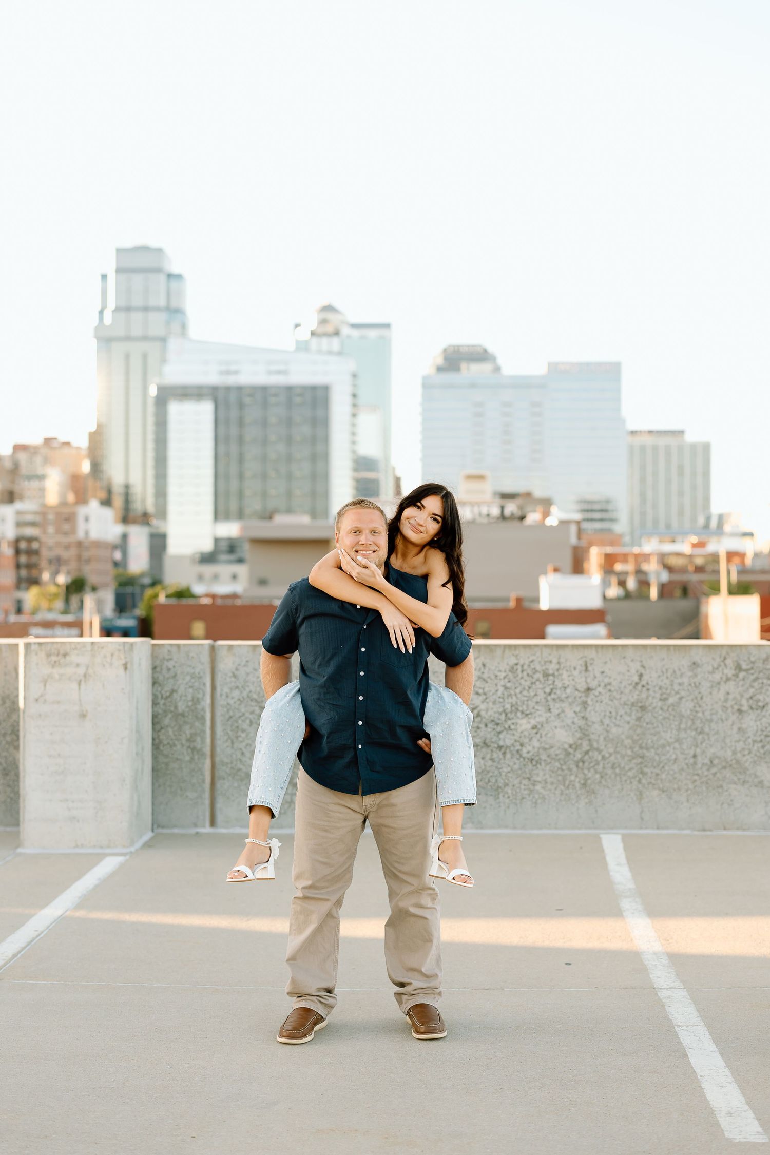 A couple poses playfully on a parking garage rooftop with the Minneapolis skyline visible in the background at sunset.