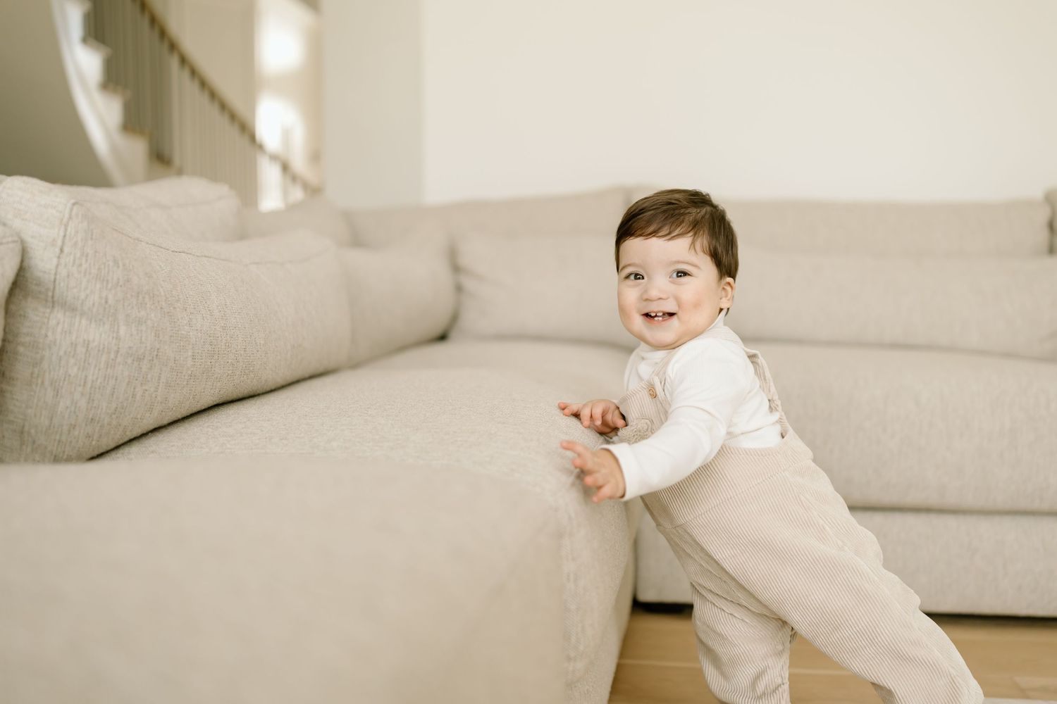 A baby in white clothing crawls and plays on a light gray couch while exploring their surroundings.