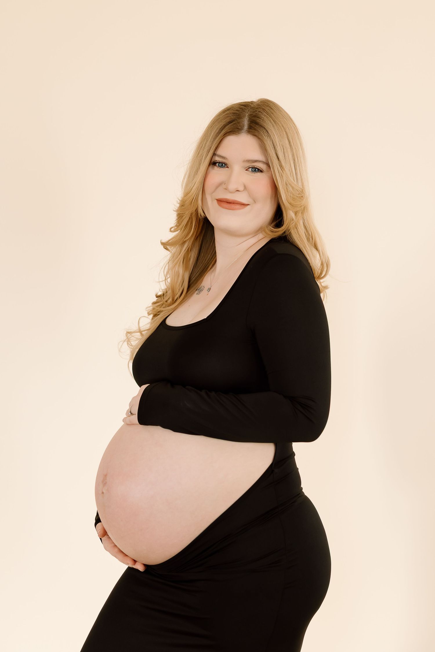 A pregnant woman in a black dress poses against a beige backdrop during a maternity photoshoot.