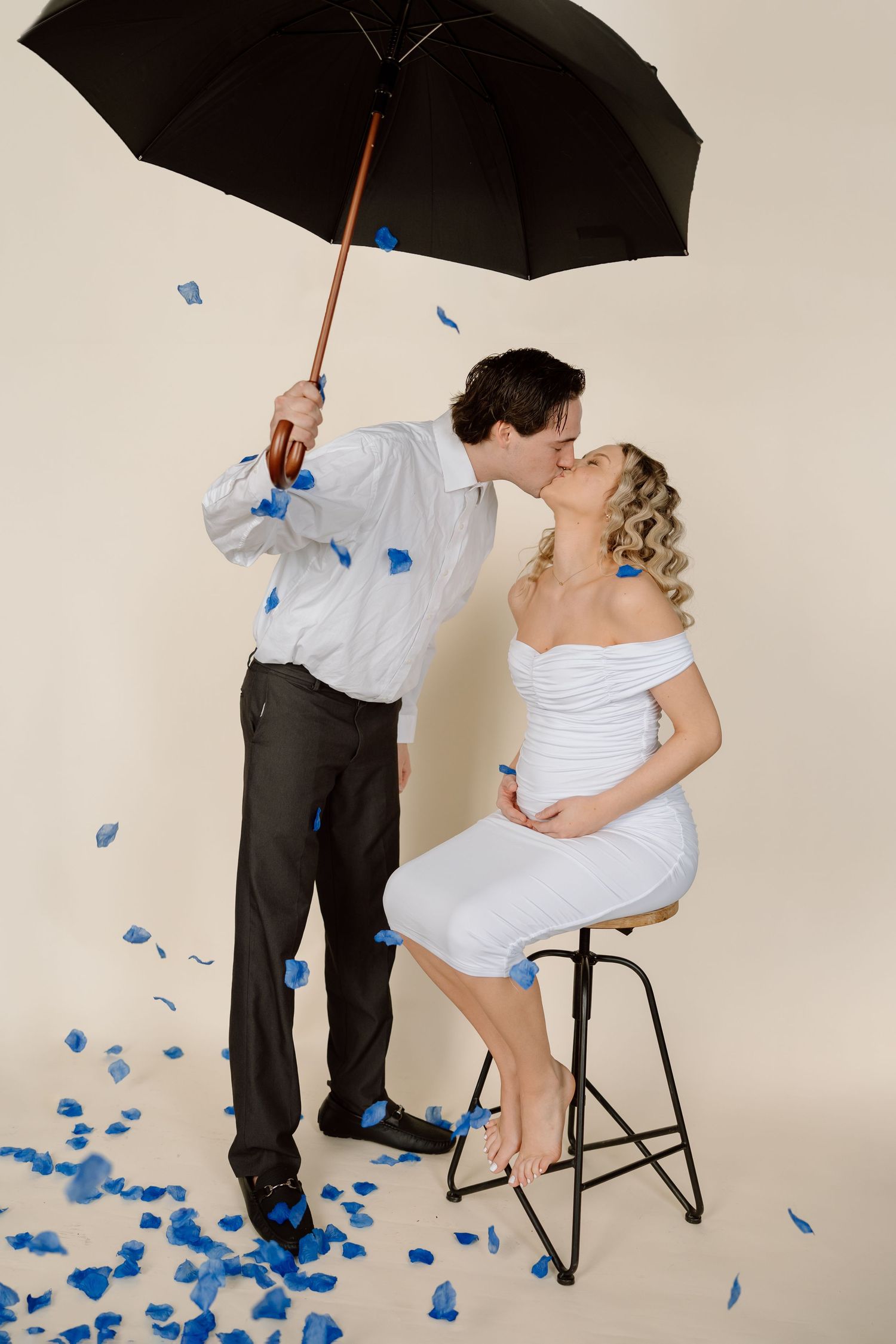 A romantic couple shares a kiss under a black umbrella with blue rose petals scattered on the ground in a photo series.