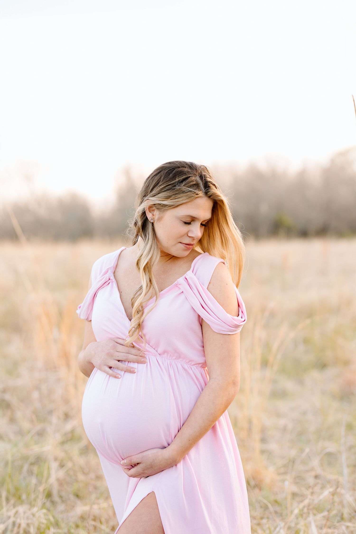 Expectant mother in soft pink dress stands in golden field during sunset while gently touching her baby bump.